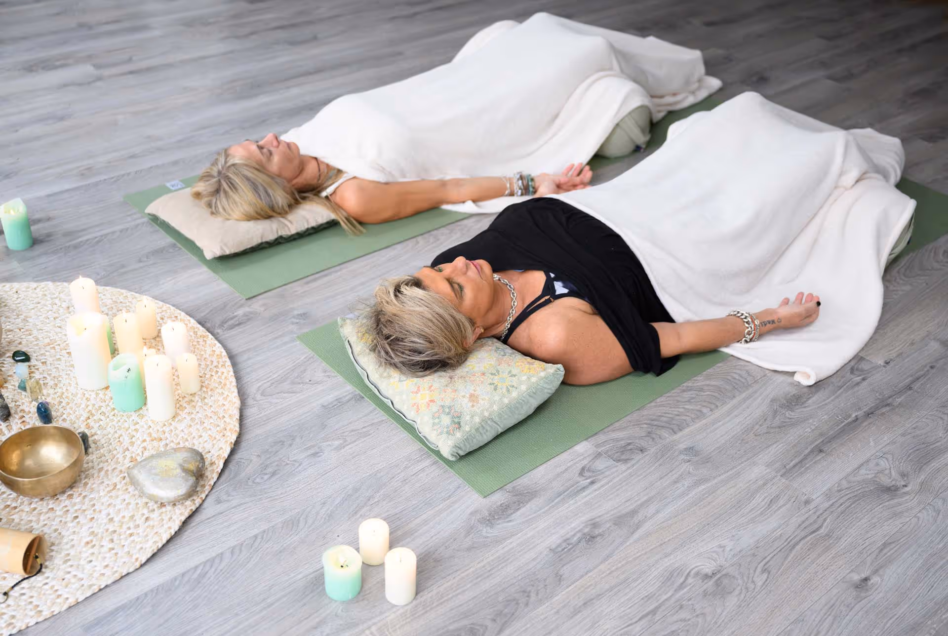 Two people lie on yoga mats with blankets, surrounded by candles on a wooden floor.