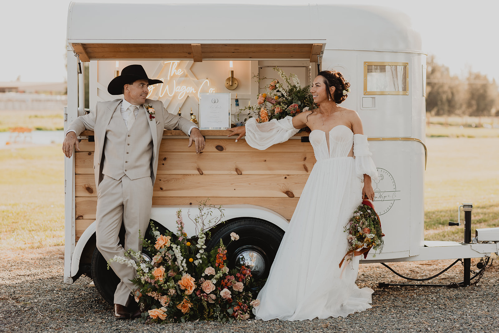 Bride in a white gown holding a bouquet and groom in a beige suit with a black cowboy hat lean on a wooden wagon bar decorated with flowers.