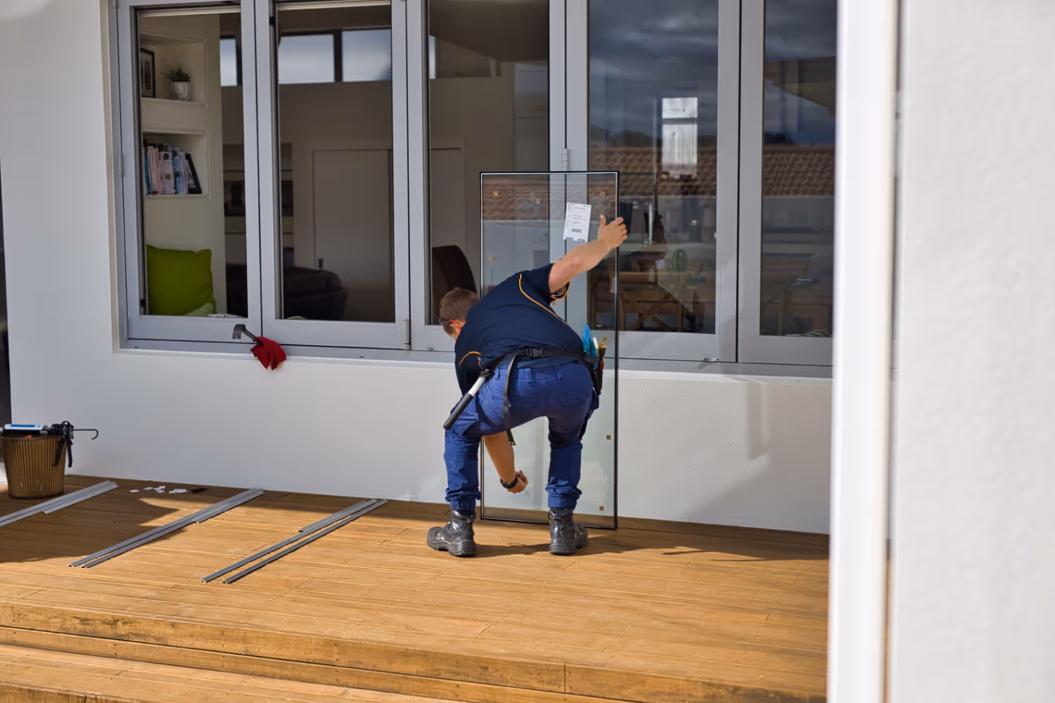 A worker in navy blue uniform installs or adjusts a glass panel on a wooden deck in front of a modern building window.