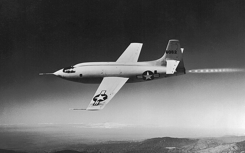 A black and white still of an aircraft in flight.