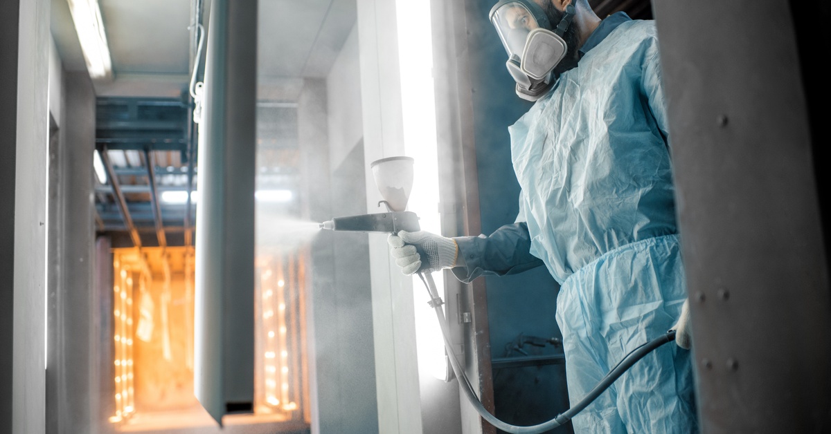 A worker in a respirator and protective suit powder coating a metal part near a high-heat industrial oven chamber.