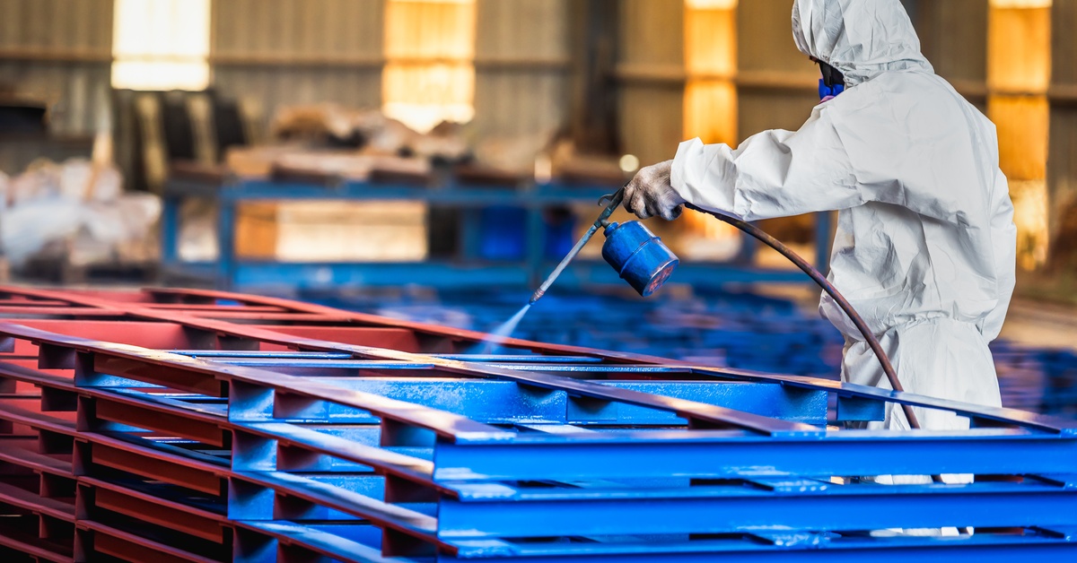 A worker in a protective suit spray coating stacked metal frames with blue and red industrial paint in a factory.