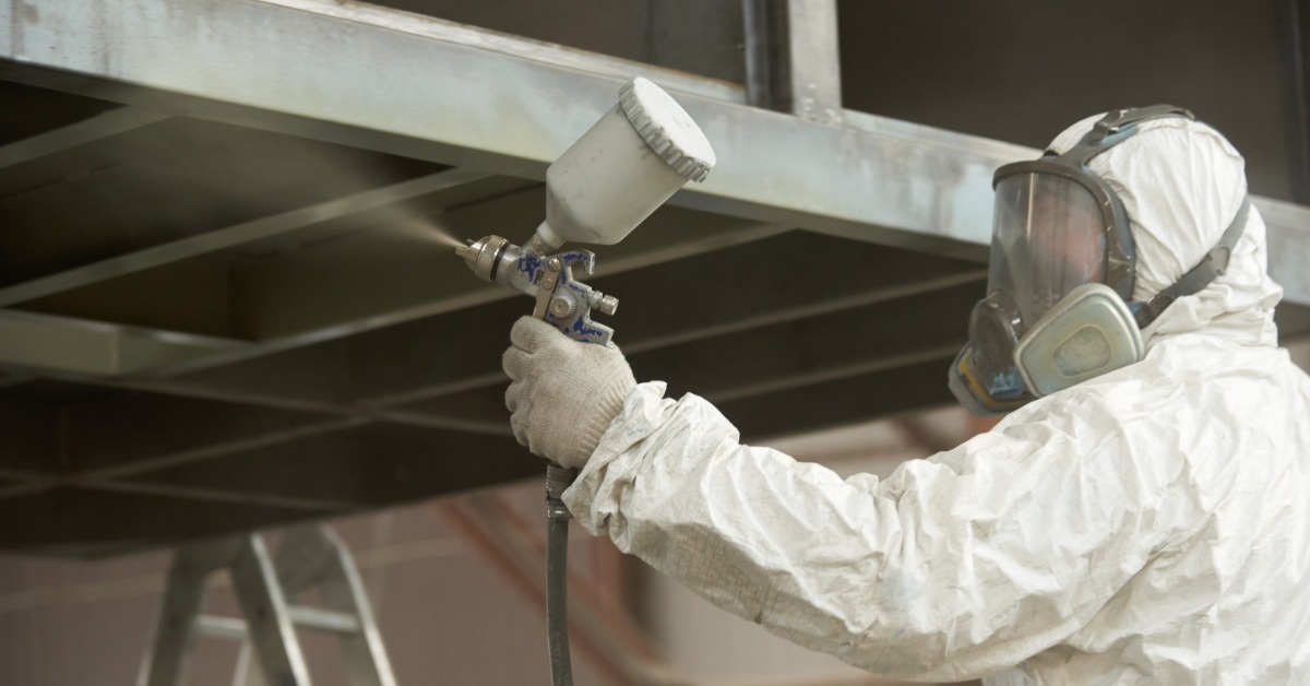 A technician wearing a protective suit and a respirator uses a spray gun to apply an industrial coating to steel beams.