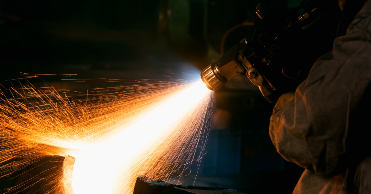 A worker uses a thermal spray torch to apply a coating to a metal part, producing a bright flame and sparks.