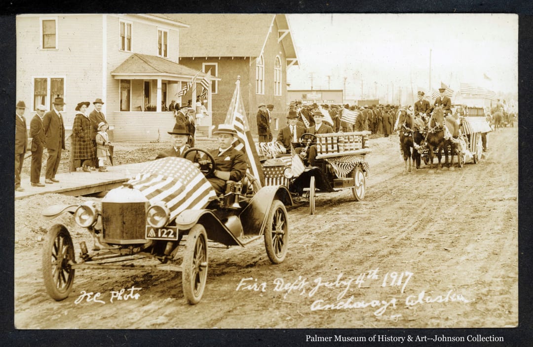This tinted photo features three vehicles in an Anchorage fourth-of-July parade in 1917.   Coming down the finely textured street toward the left of the camera, the first car in left foreground is a roadster containing two men in suits, flying the American flag from the stern with a duplicate flag covering the engine hood.    A second car, mostly covered in bunting, follows, also containing two men in suits.  In far middle ground at right two men in firemen uniforms drive two horses pulling an apparent fire apparatus covered in bunting and flying flags.  Beyond the cars at left foreground spectators stand on the sidewalk.  At left middle ground stand a two-story white house and a large building resembling a church.  Beyond the church a large crowd is assembled along the sidewalk.  Beyond the crowd past the church several other buildings are visible, one displaying a sign proclaiming the "Anchorage Daily Times".  In background at extreme right a man on horesback and many American flags are visible.