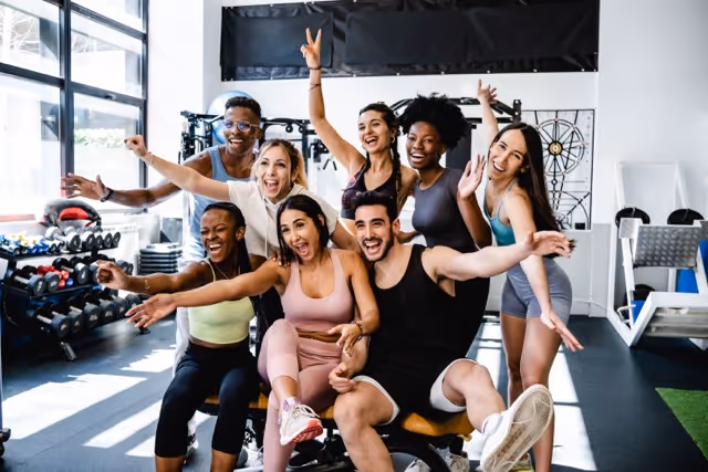 Un grupo diverso y alegre de personas en un gimnasio, celebrando y posando para la cámara. Están vestidos con ropa deportiva y están dispersos alrededor de una bicicleta estática, algunos están sentados en ella y otros están de pie. Sus expresiones y gestos sugieren que se lo están pasando muy bien, y algunos levantan las manos en alto y sonríen ampliamente. El fondo muestra equipos de ejercicio, como pesas libres, lo que sugiere un ambiente de entrenamiento alegre y comunitario.