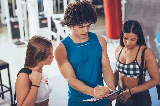 Una instructora de acondicionamiento físico con cabello rizado y vestida con una camiseta azul sin mangas, sostiene un portapapeles y discute con dos atentas clientas en un gimnasio. Las mujeres están vestidas con ropa deportiva, lo que indica una interacción profesional, posiblemente con respecto a sus planes de acondicionamiento físico o su progreso. El trasfondo está desenfocado, lo que sugiere la naturaleza privada y enfocada de la conversación.