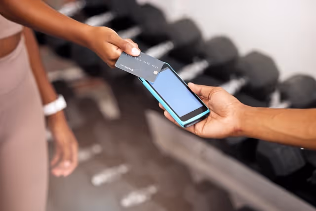 Un primer plano de dos personas en un gimnasio intercambiando una tarjeta de crédito y un teléfono móvil, presumiblemente procesando un pago. La persona que entrega la tarjeta de crédito lleva un vestido o una falda de color claro y tiene un rastreador de actividad física o una pulsera en la muñeca, lo que sugiere que es un cliente. La otra persona sostiene el teléfono, que está envuelto en una funda protectora azul, lo que indica que se está realizando una transacción. En el fondo suavemente enfocado, se ven montones de mancuernas, lo que indica que el escenario es un gimnasio.