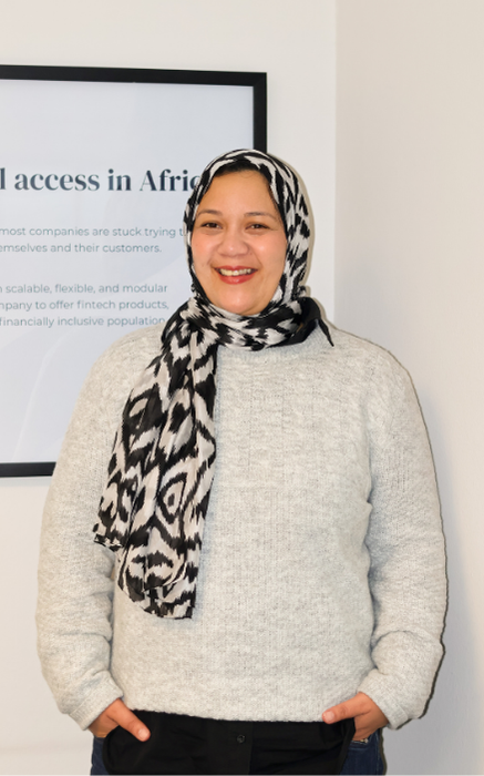 Smiling woman wearing a black and white patterned hijab and a light gray sweater standing with hands in pockets in front of a framed poster on a white wall.