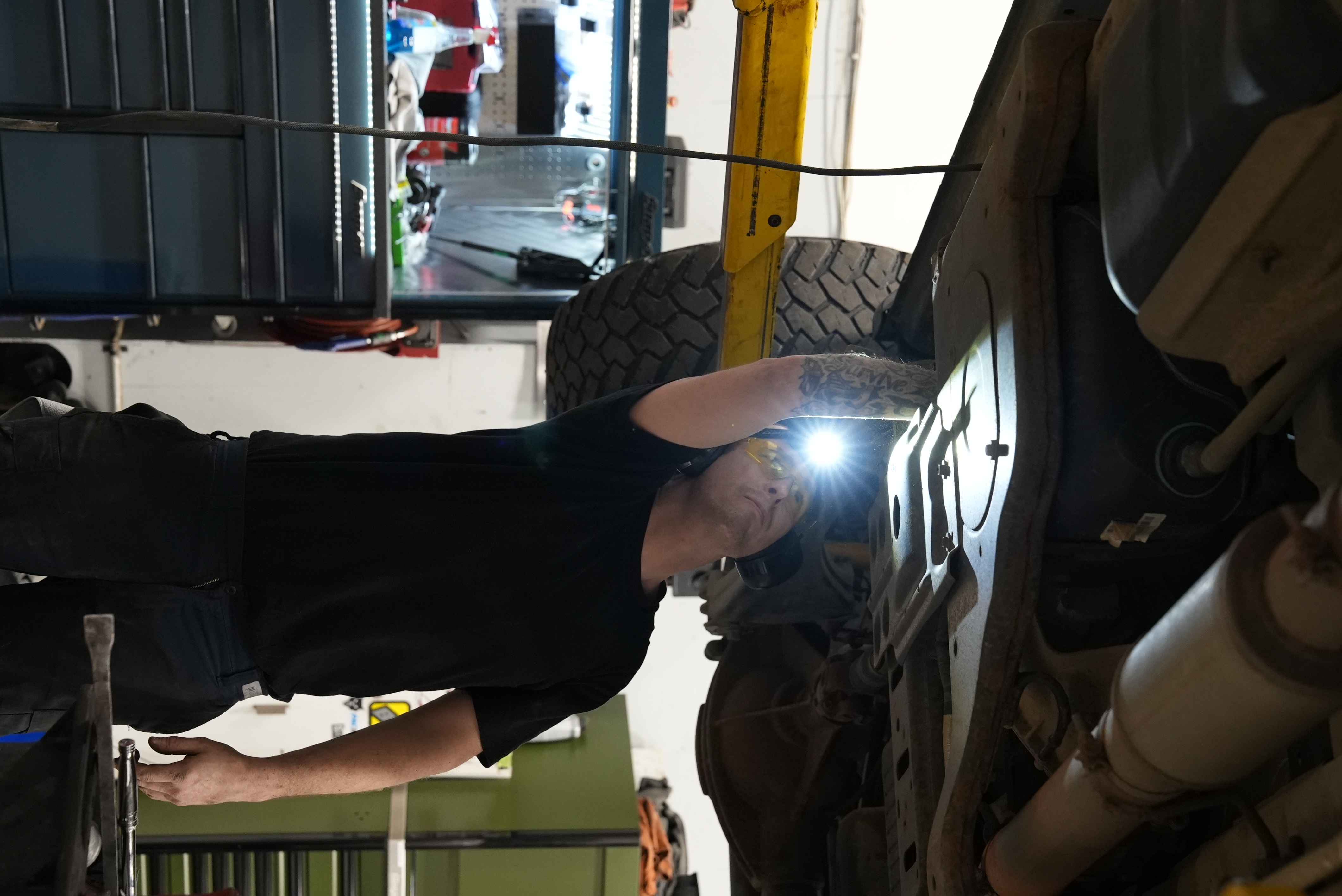 Mechanic wearing safety glasses and ear protection inspecting the underside of a vehicle in a garage.