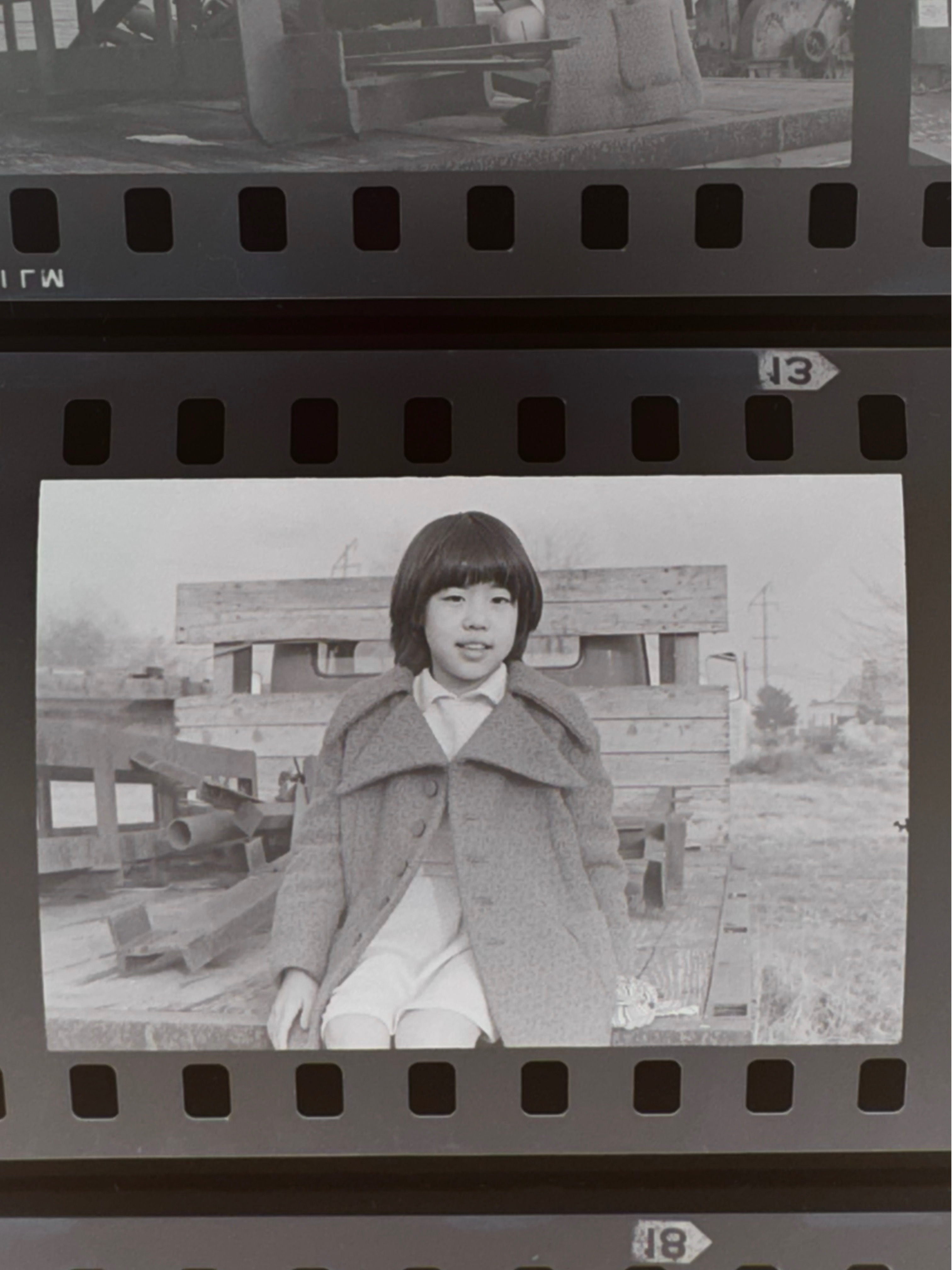 Kelly Goto as Chieko — a young girl with a bowl cut in a grey coat and white knee socks, smiling outdoors. Gold Watch, 1977. Photo by Steve Suzuki.