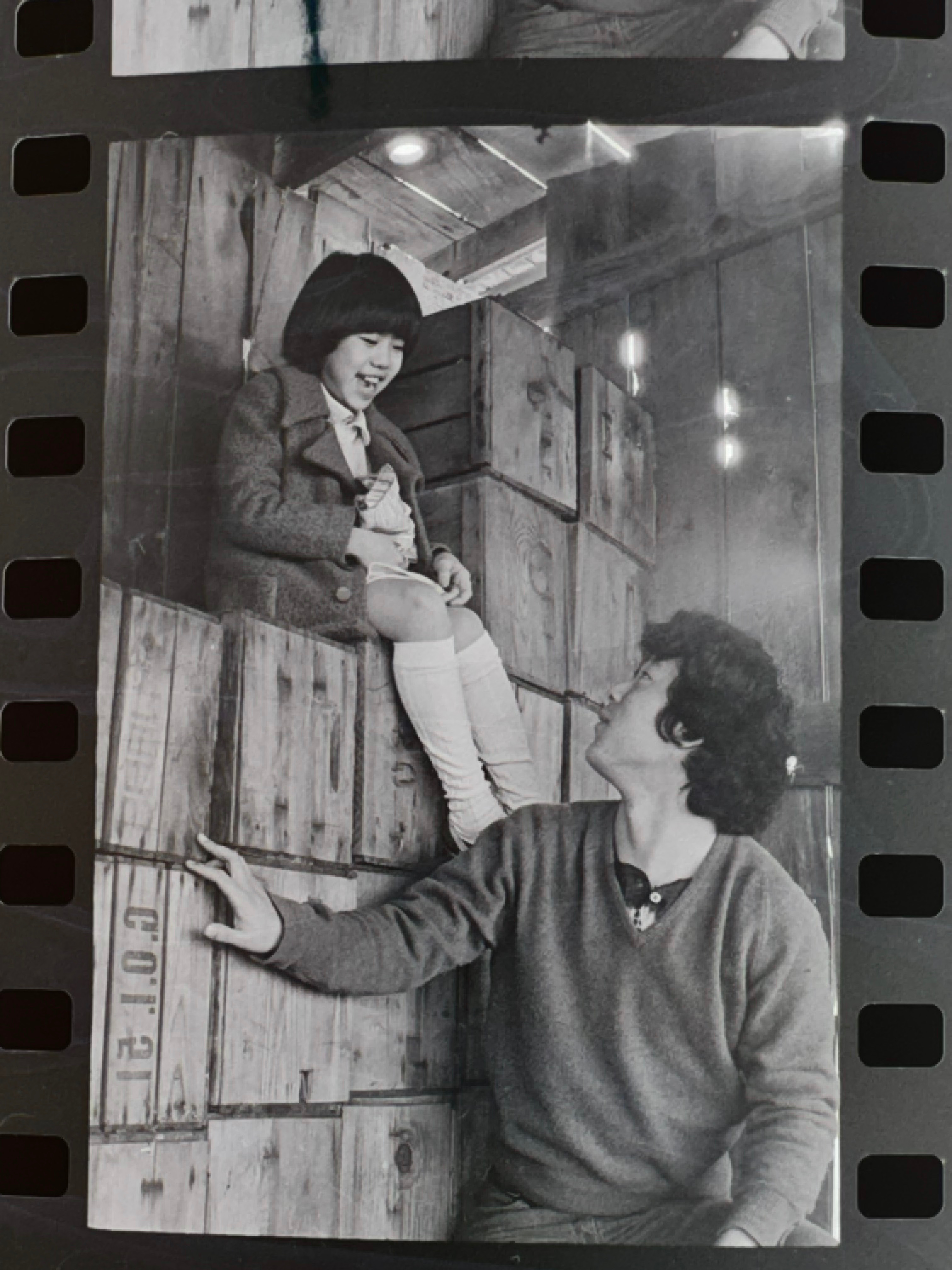 A young girl in a grey coat laughs while perched atop wooden apple crates, as an adult woman looks up at her warmly. Gold Watch, 1977. Photo by Steve Suzuki.