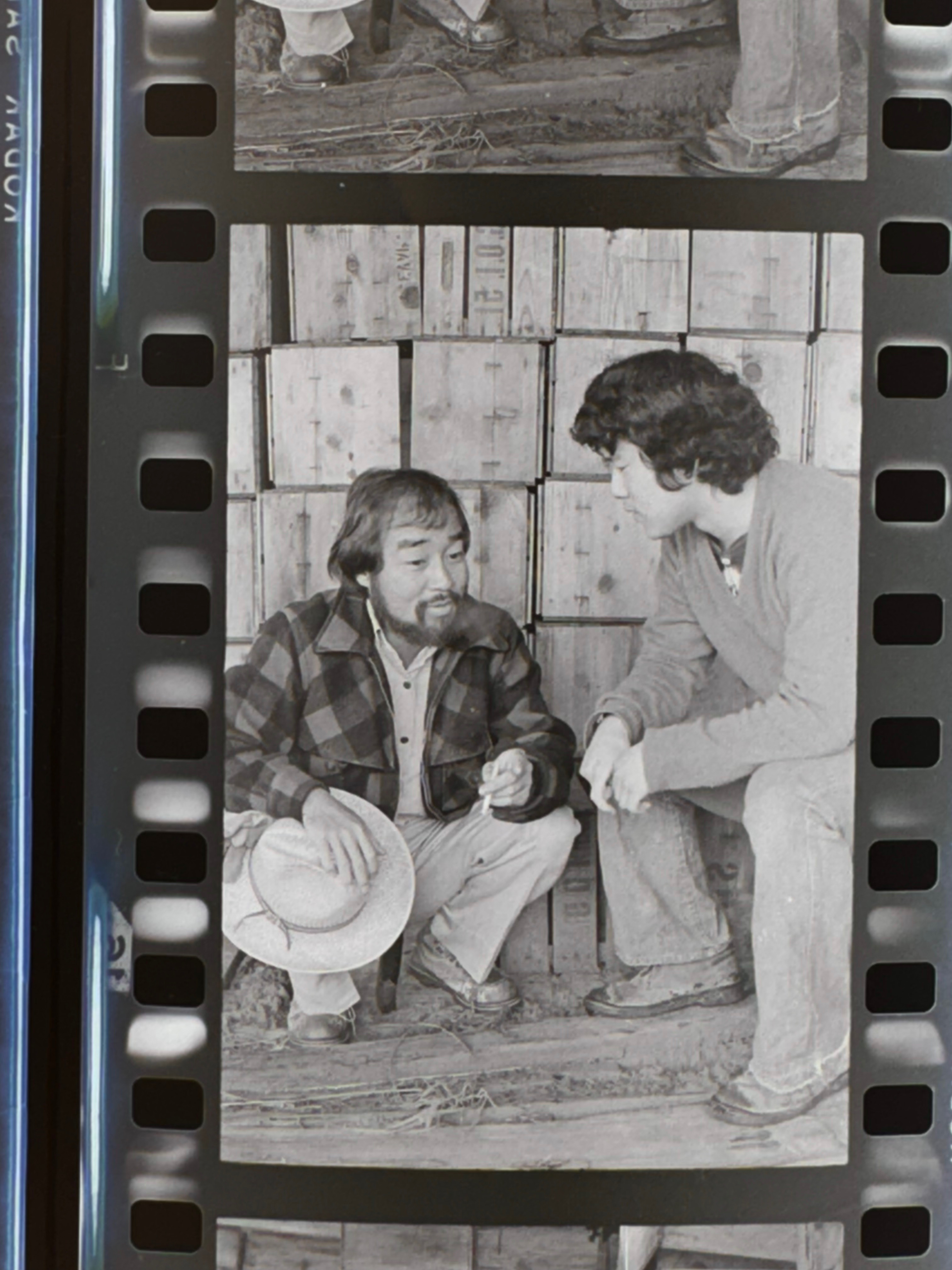 Two men in conversation seated against a wall of wooden apple crates, one holding a straw hat. Gold Watch, 1977. Photo by Steve Suzuki.