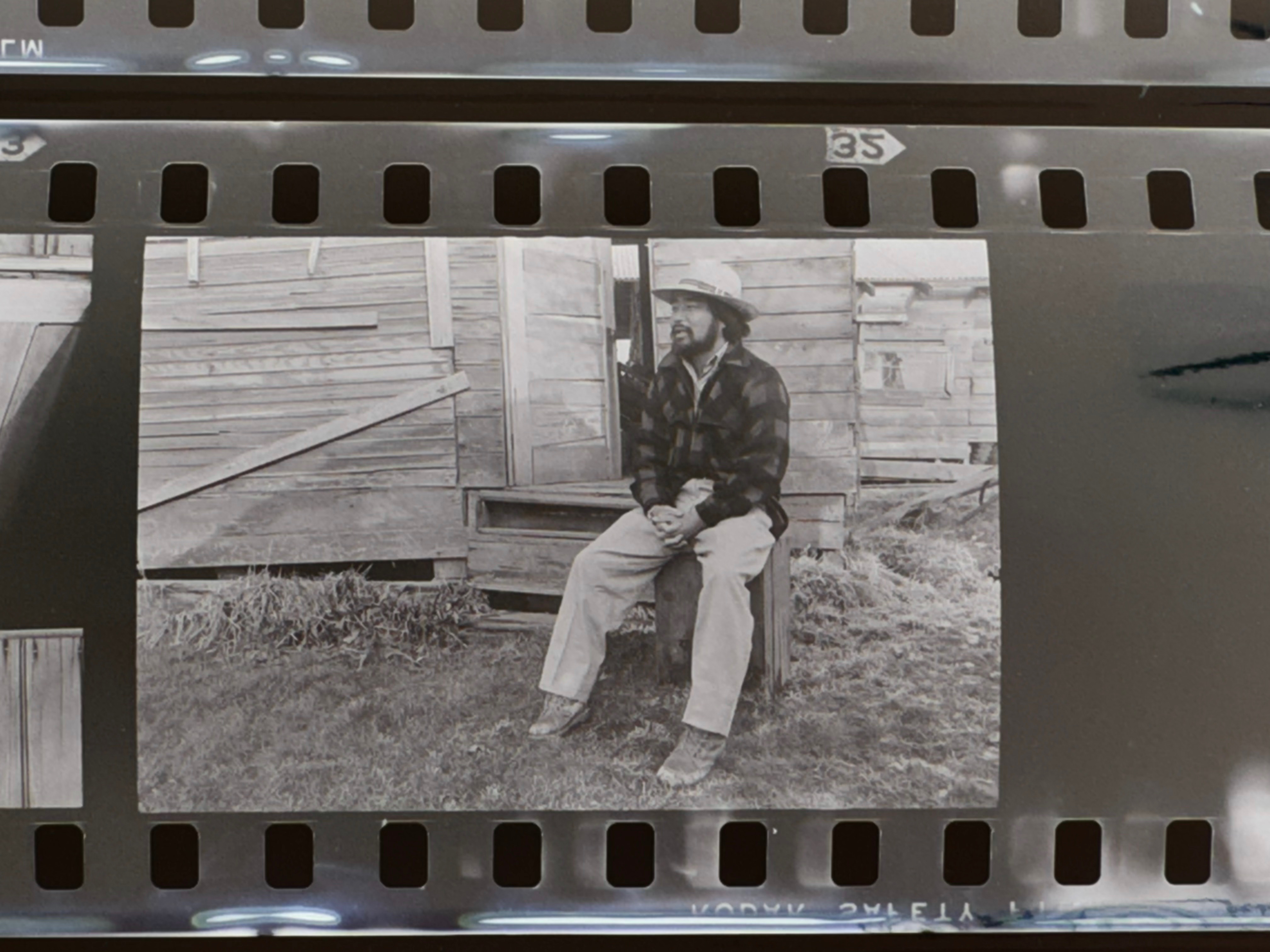 A man in a plaid jacket and straw hat sits alone on the steps of a weathered farmhouse, hands clasped, gazing into the distance. Gold Watch, 1977. Photo by Steve Suzuki.
