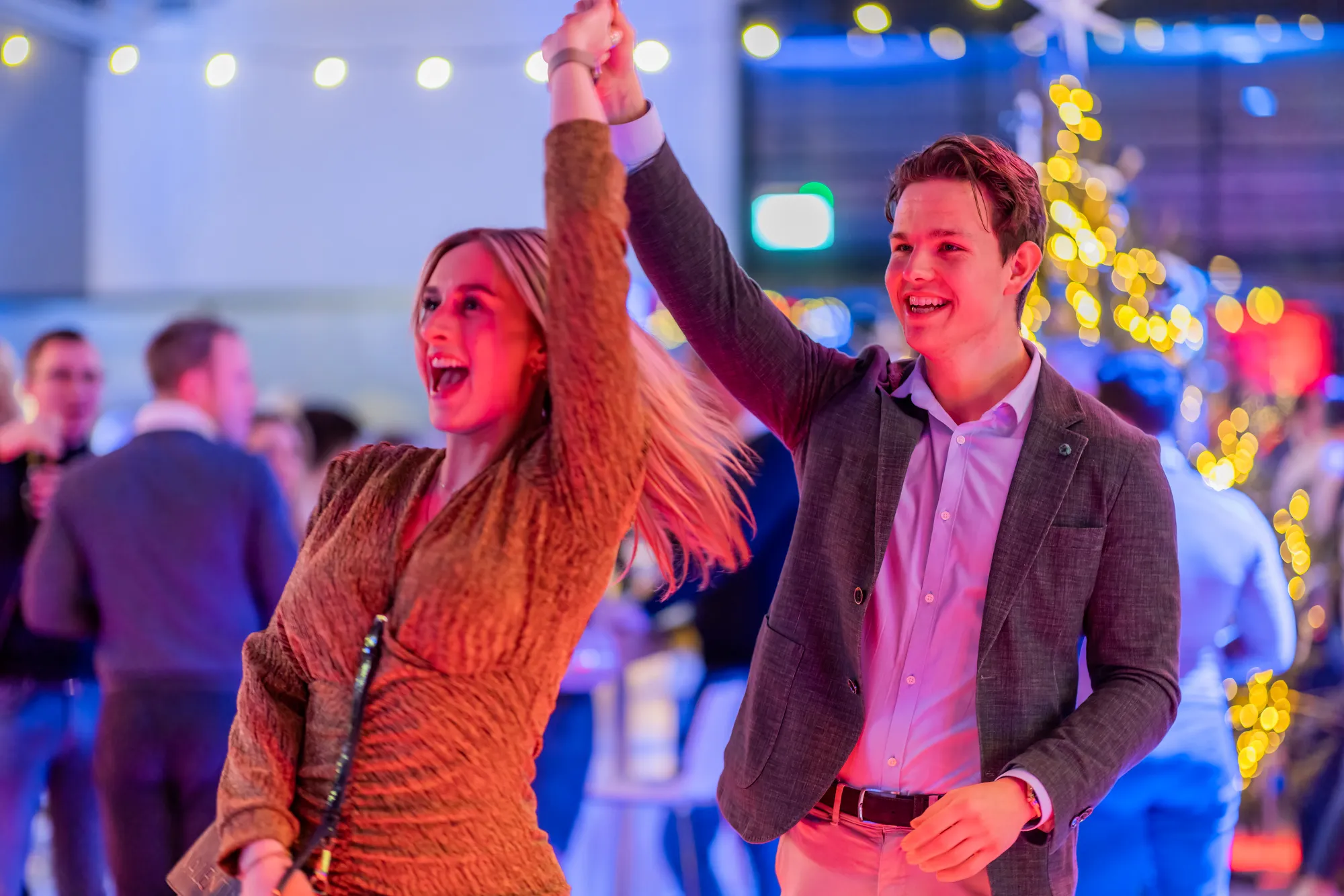 Young man and woman smiling and dancing together at a lively social event with colorful lights and bokeh in the background.