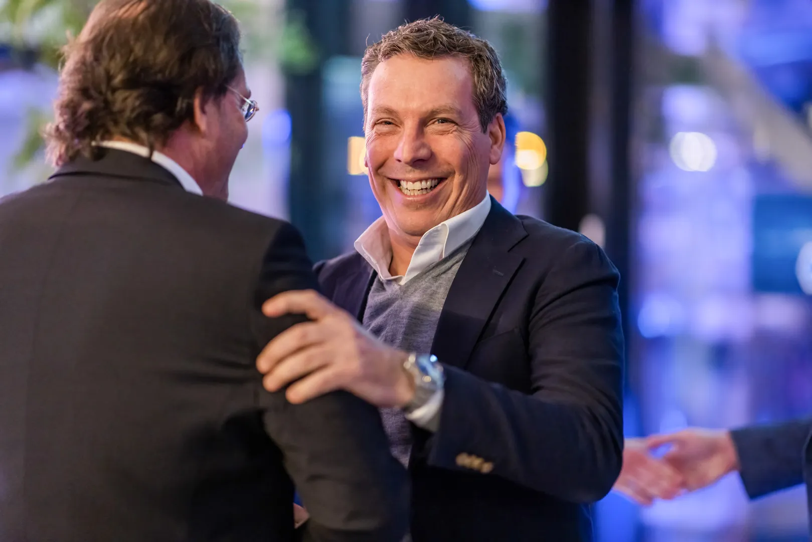 A smiling man in a blazer and sweater warmly greets another man in a suit during a casual business event.
