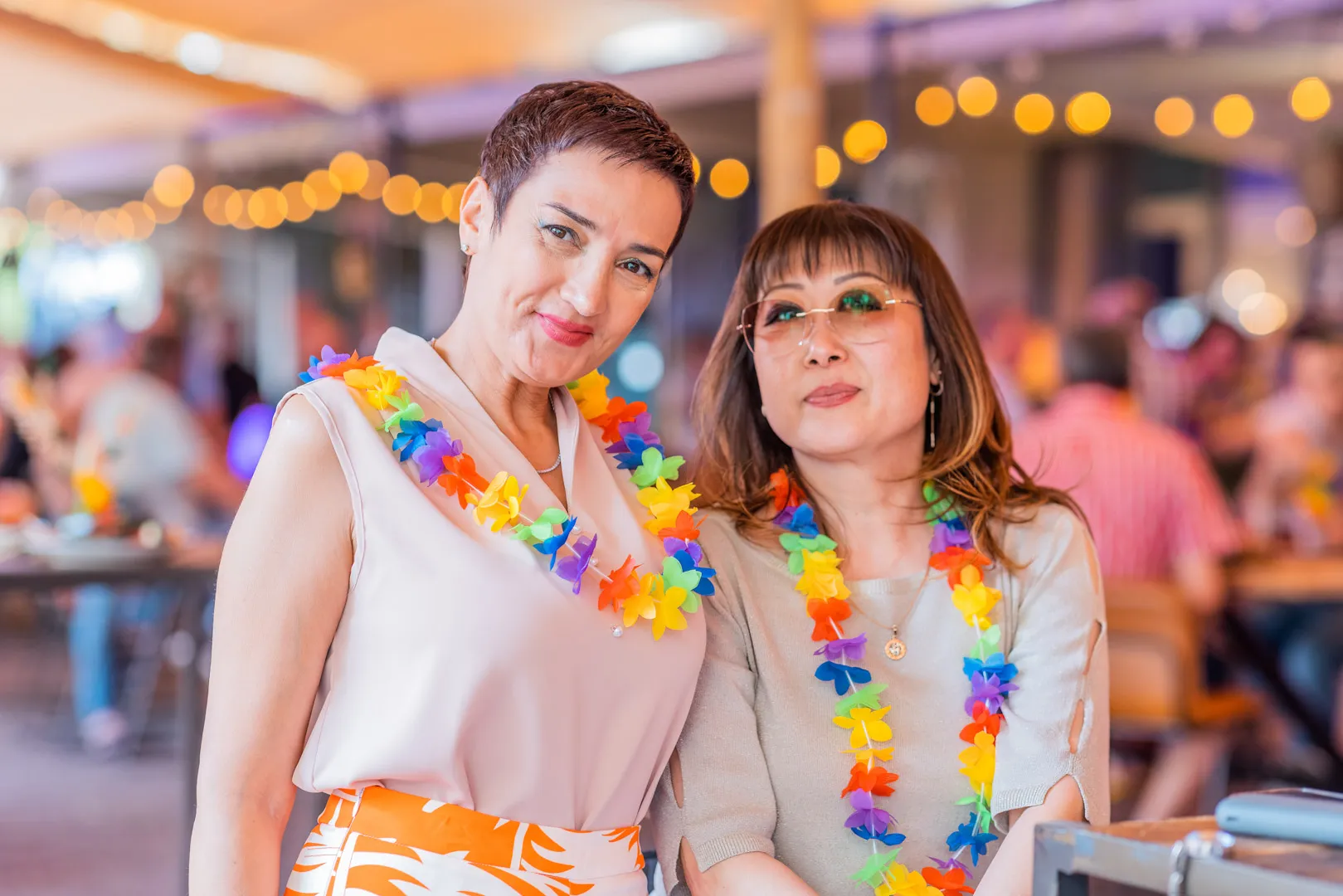 Two women wearing colorful flower leis posing together at a lively indoor event with string lights in the background.