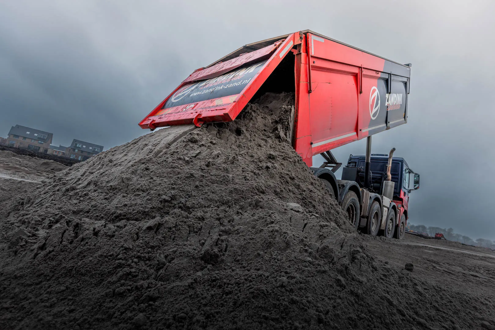 Red dump truck unloading a large pile of sand or soil on a cloudy day at a construction site.