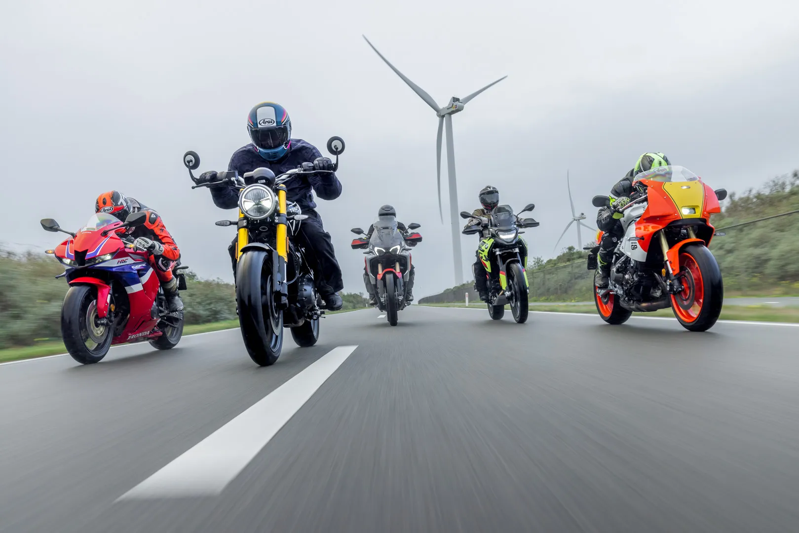 Five motorcyclists riding on a road with wind turbines in the background under cloudy sky.