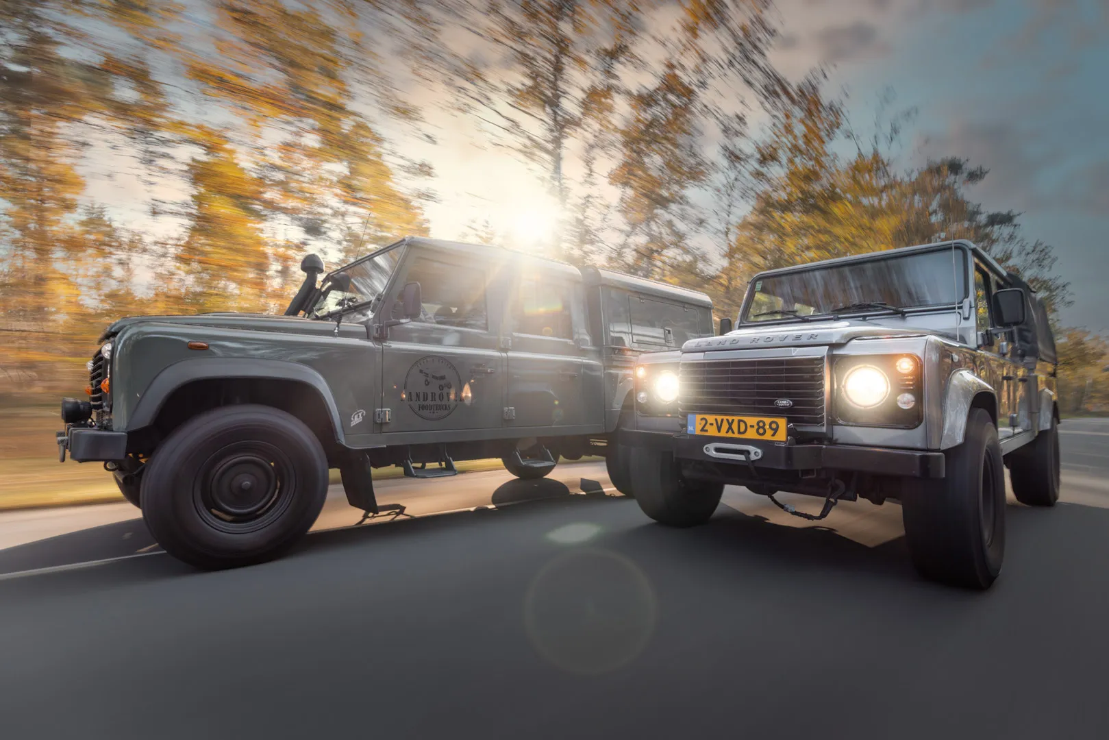 Two Land Rover Defender SUVs driving side by side on a road with autumn trees and sun flare in the background.