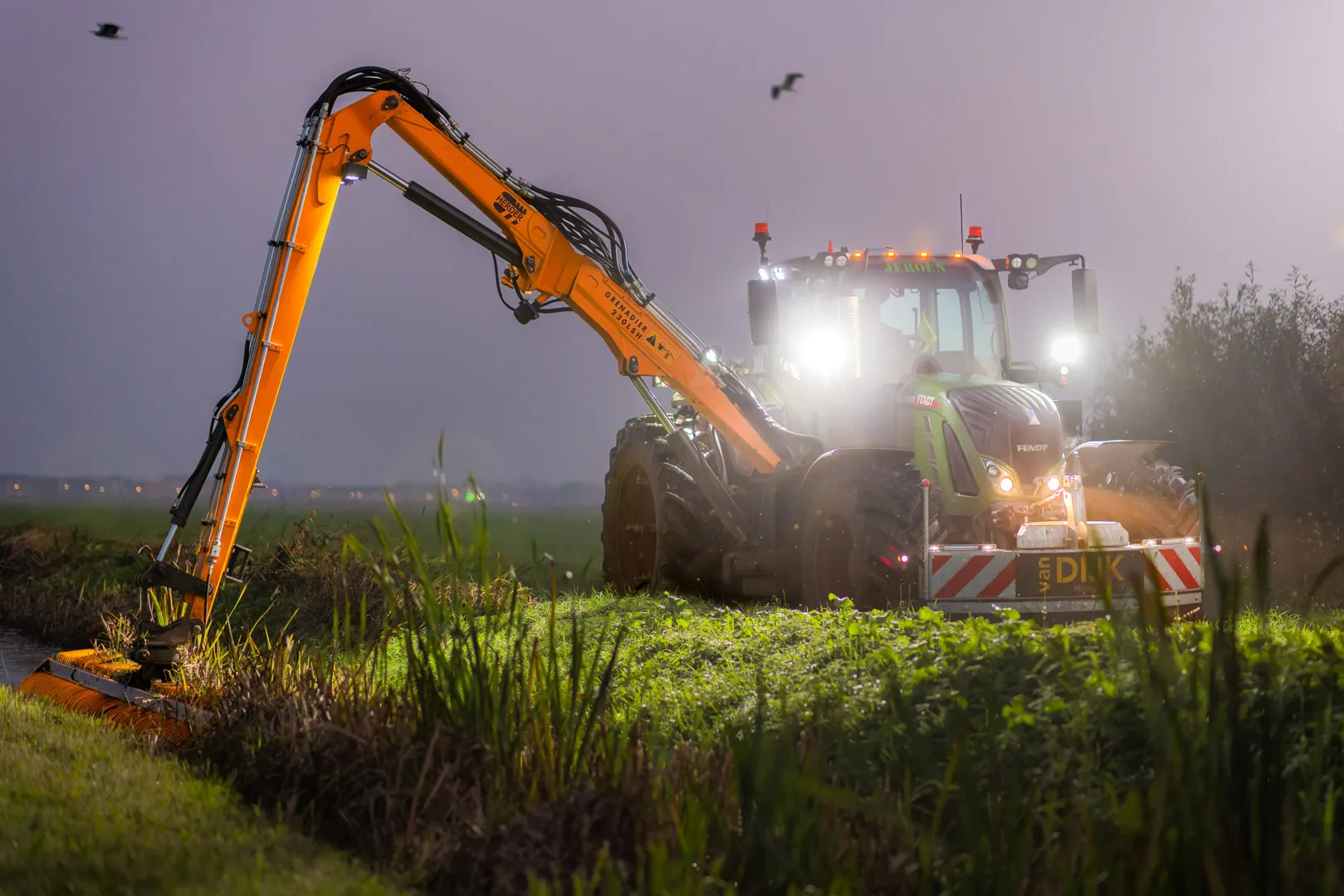 Fendt tractor with long orange arm mowing grass by a roadside canal at dusk with headlights on.