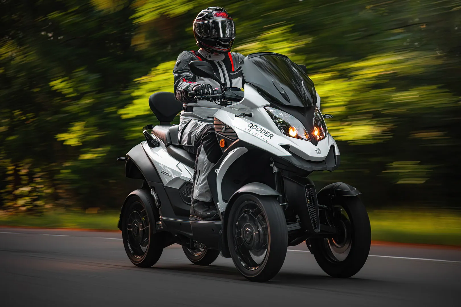 Person wearing black and gray riding gear and helmet riding a white and black four-wheeled motorcycle on a road with green forest background.