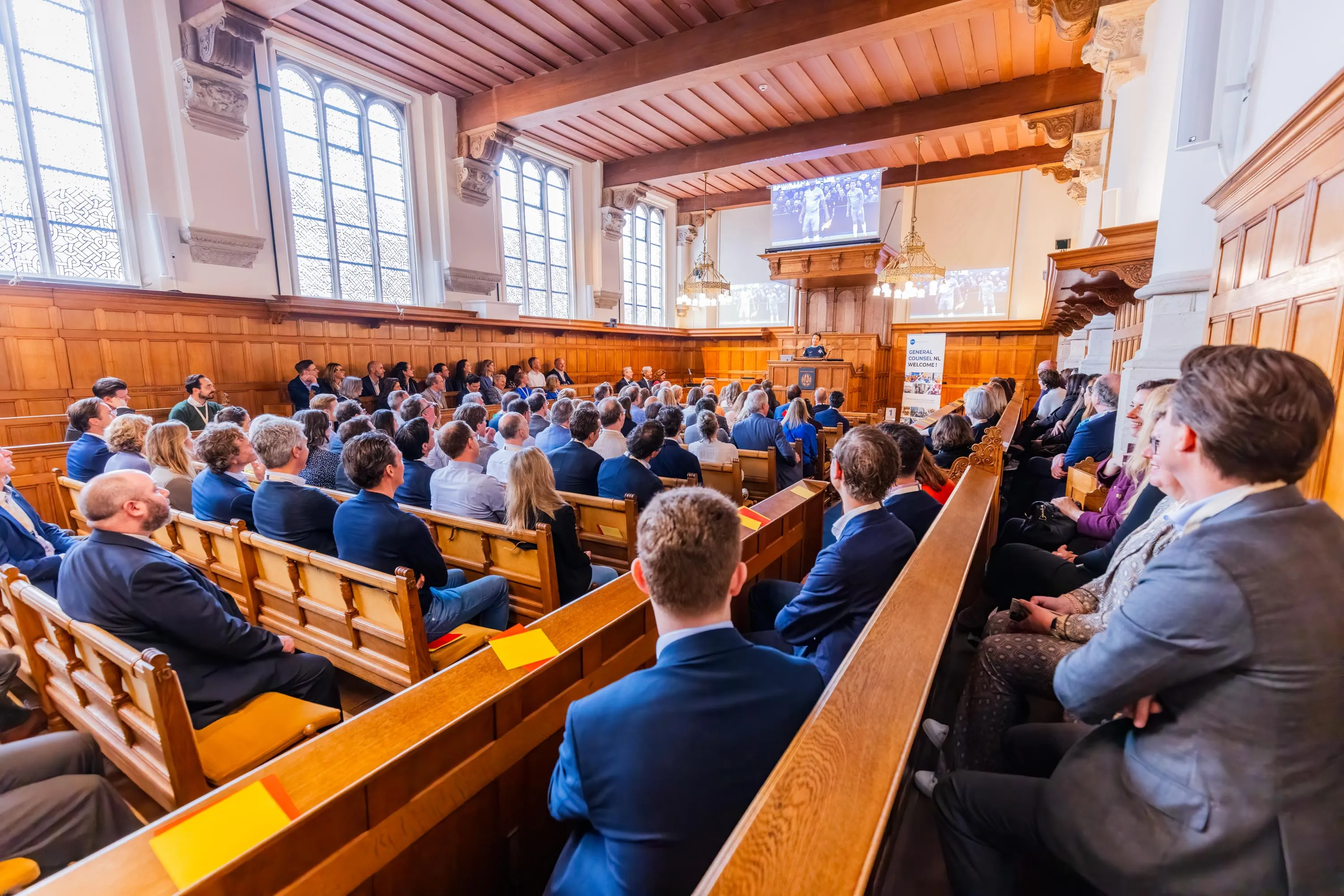 Audience of professionally dressed people seated in a wood-paneled room listening to a speaker at a podium with projected images behind.