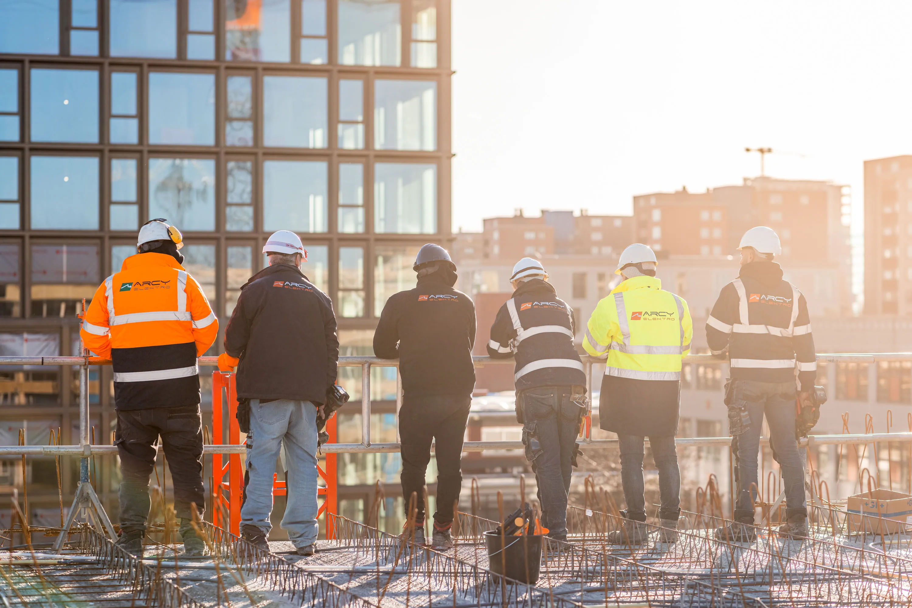Six construction workers wearing protective gear and jackets with Arcy Elektro logo standing on a building site overlooking city buildings.