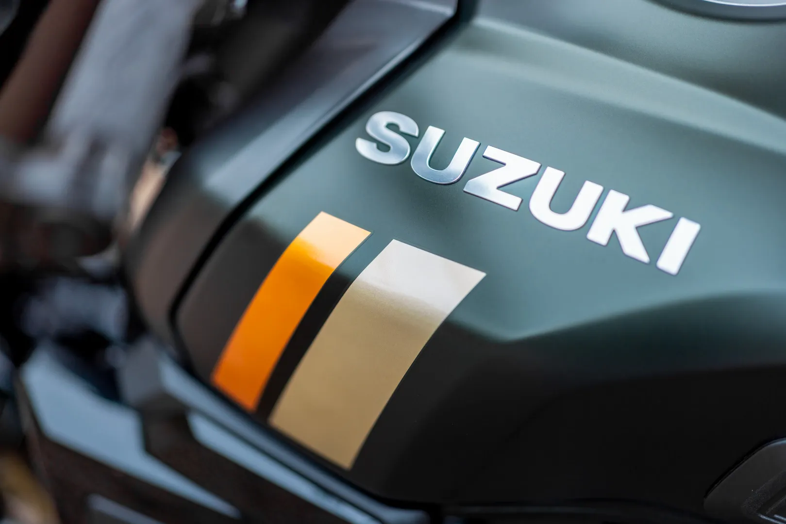 Close-up of a Suzuki motorcycle fuel tank with two vertical stripes in orange and beige.