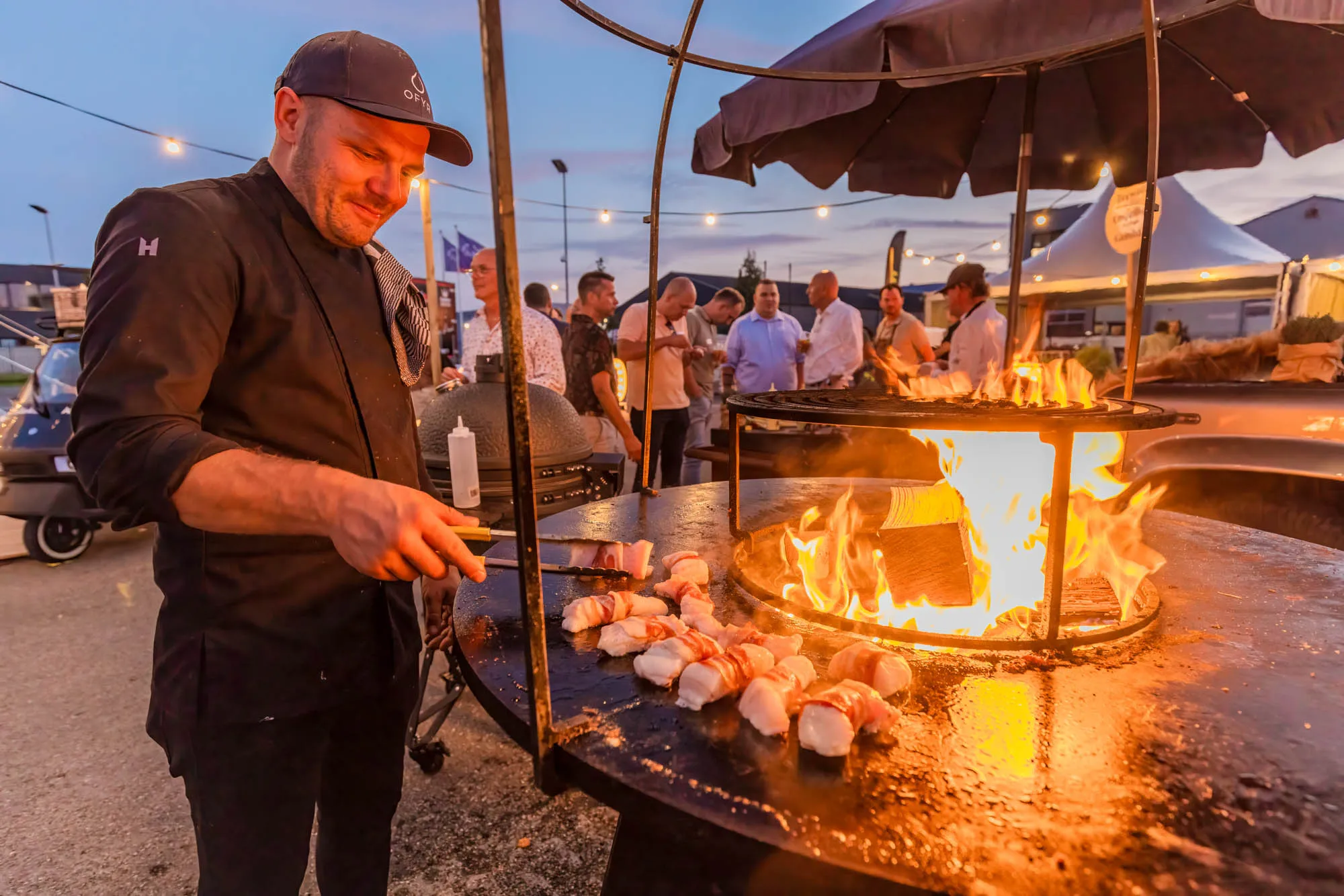 Chef cooking bacon-wrapped food on a large outdoor fire grill at an evening event with people in the background.