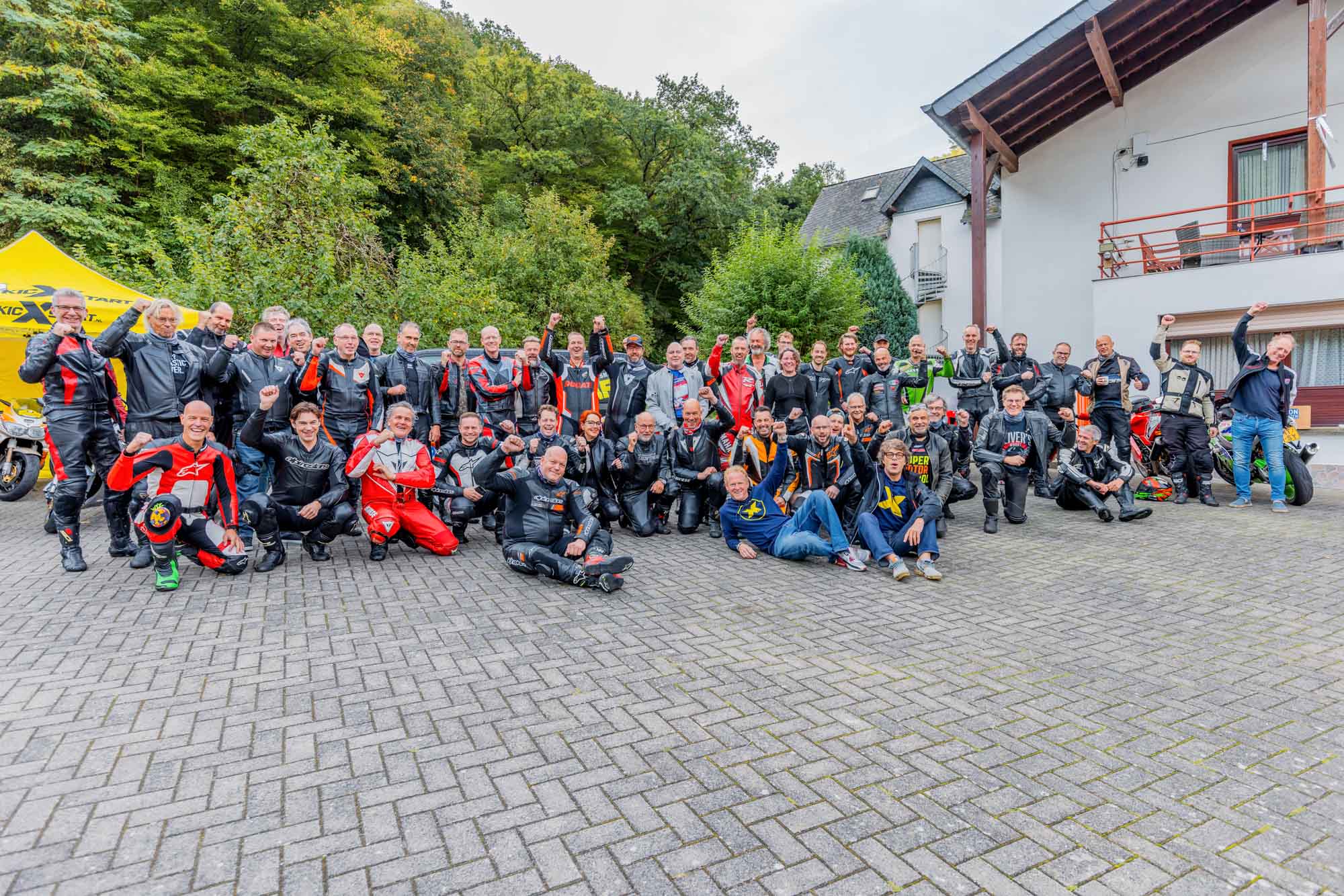 Large group of motorcyclists in riding gear posing outdoors on a paved area with trees and a building in the background.