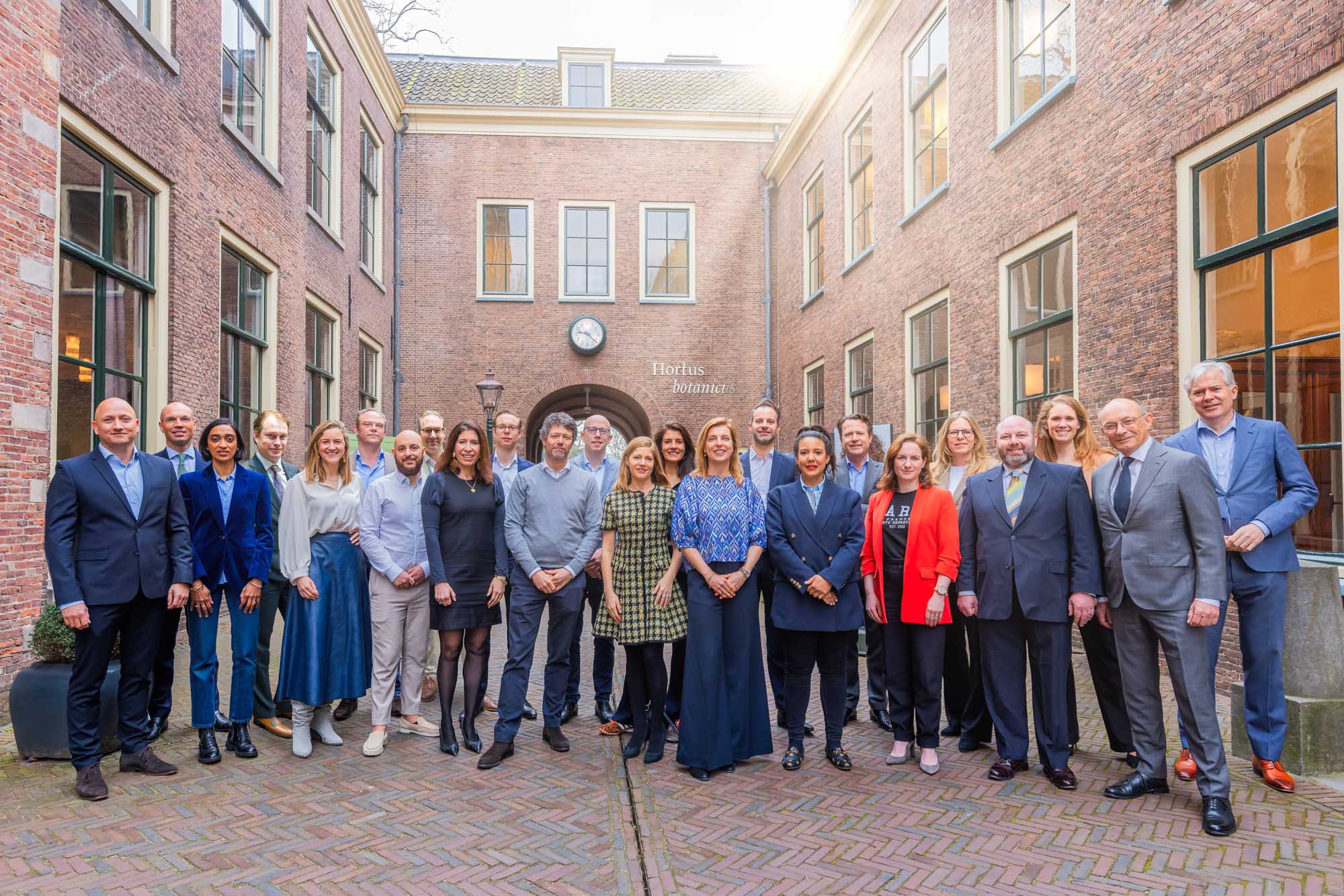 Group portrait of 22 diverse professionals standing in a brick courtyard with tall windows and a clock on the wall.