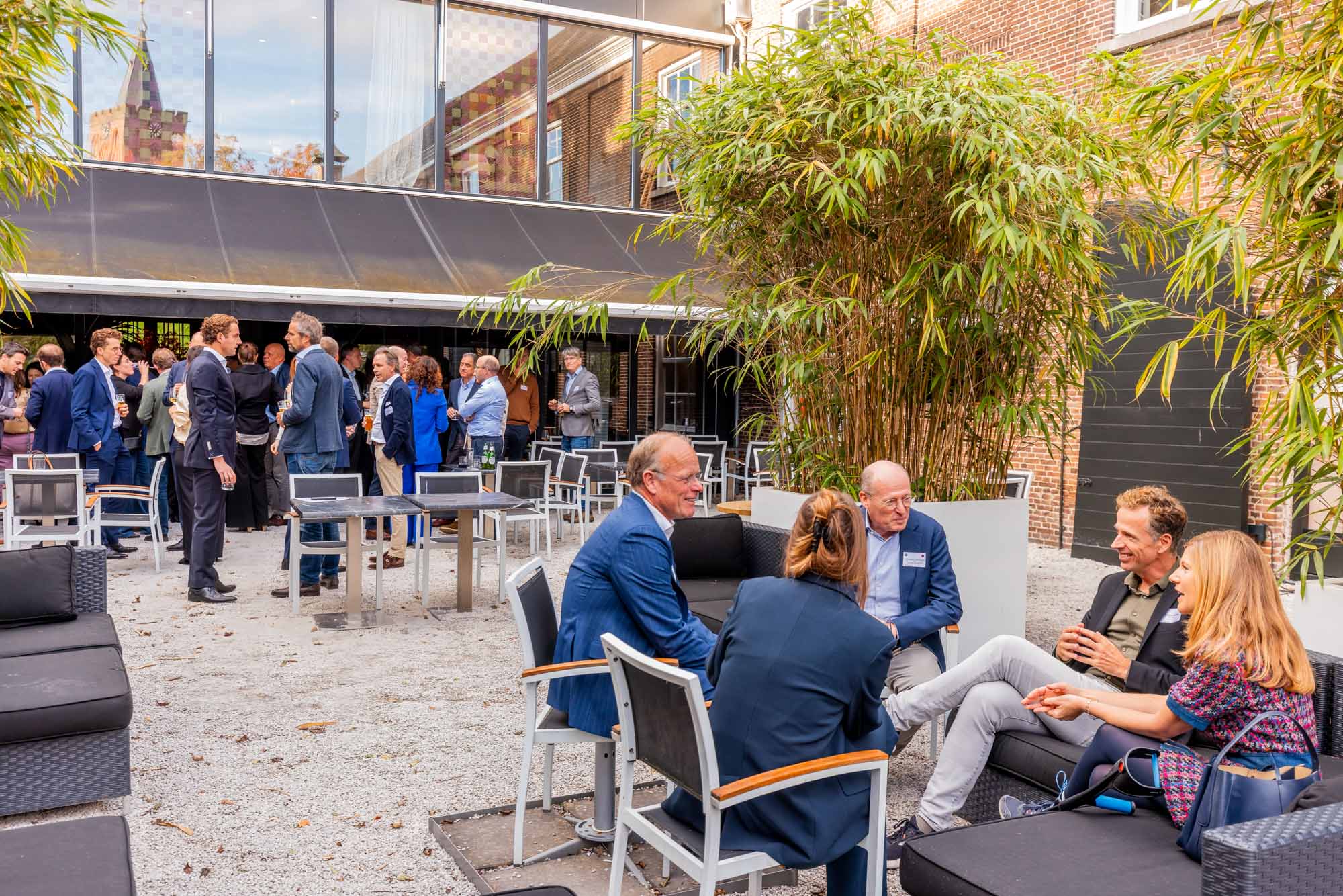 Group of people socializing at an outdoor patio event with black seating and green plants.