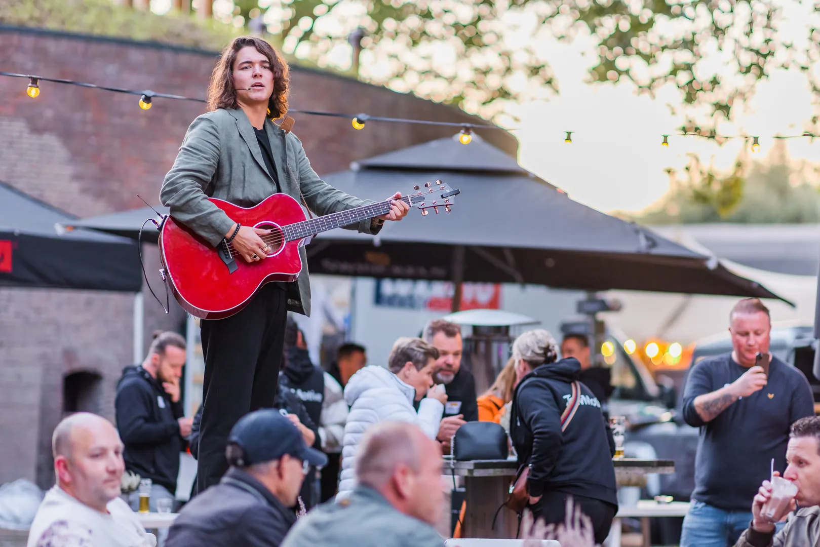 Young musician playing a red acoustic guitar and singing with a headset microphone at an outdoor event with people socializing in the background.
