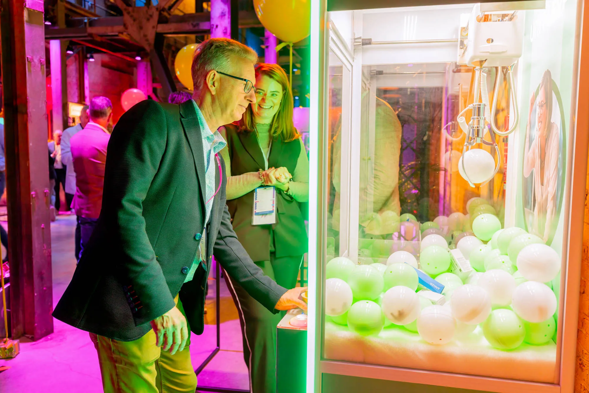 Man in glasses and blazer playing a claw machine with white balls inside, watched by a smiling woman in a vest.