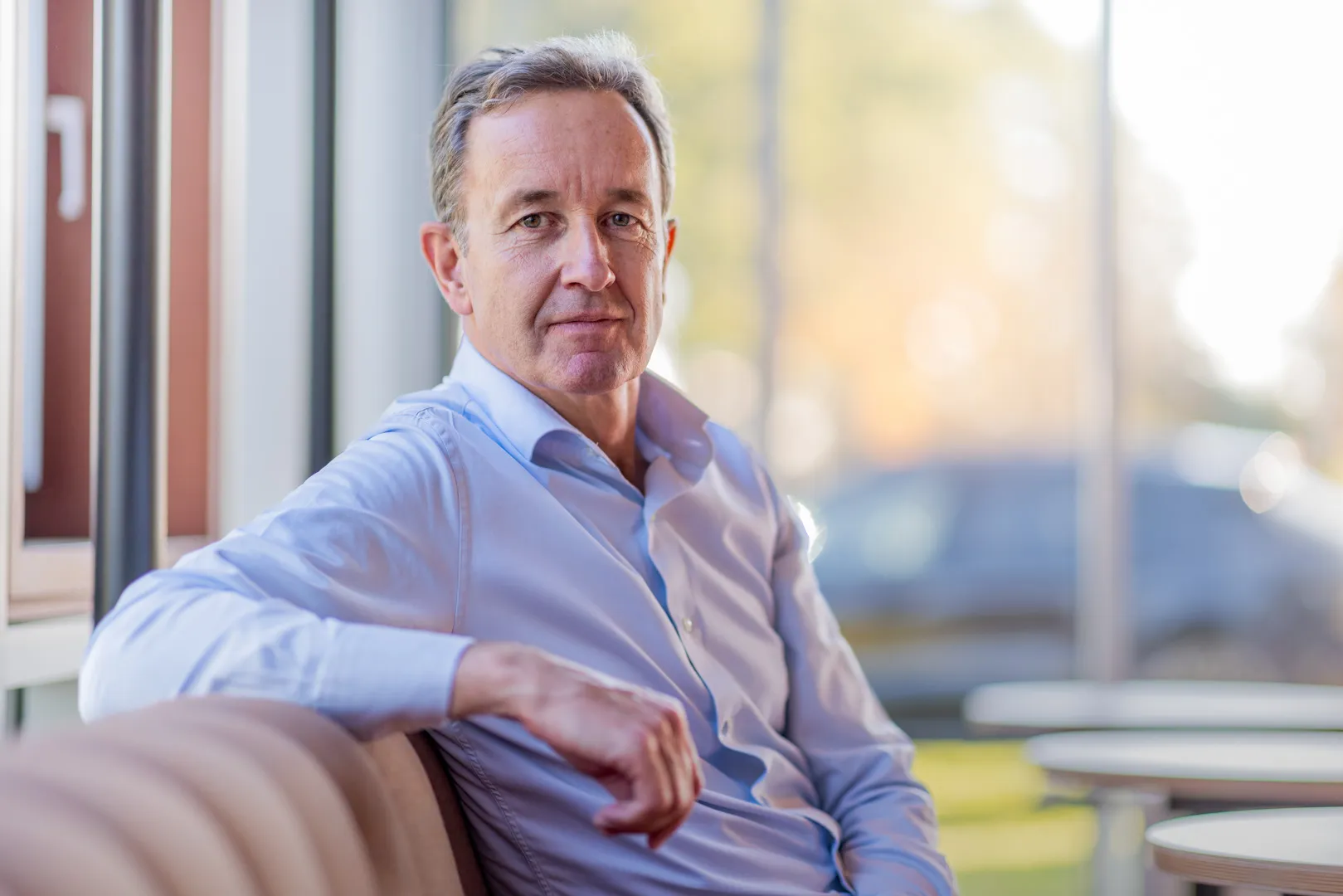 Middle-aged man in a light blue shirt sitting on a beige sofa near large windows with a blurred outdoor background.