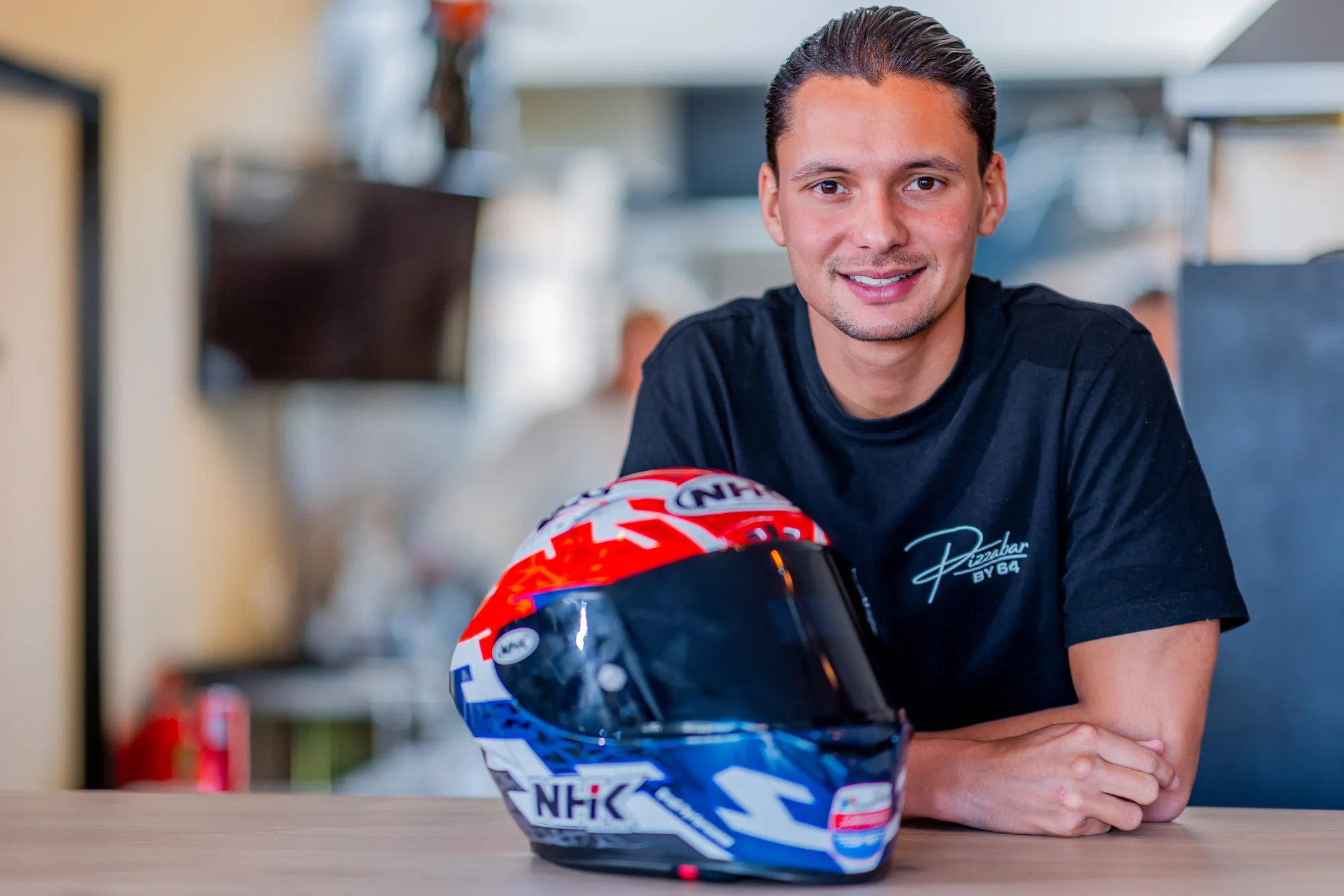 Smiling man in a black t-shirt sitting at a table with a colorful NHK motorcycle helmet in front of him.