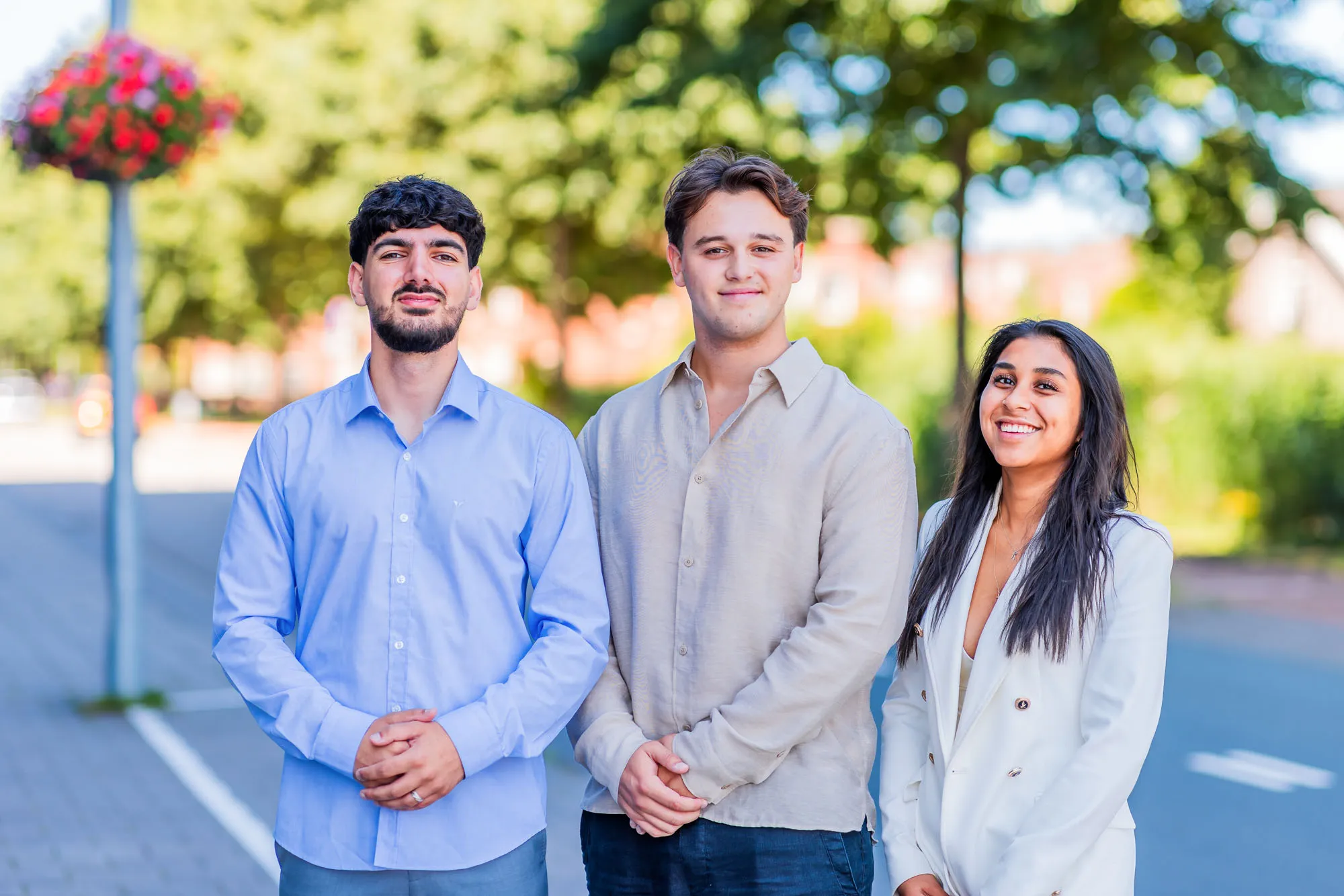 Three young adults standing outdoors on a sunny day, smiling and dressed in smart casual clothing.