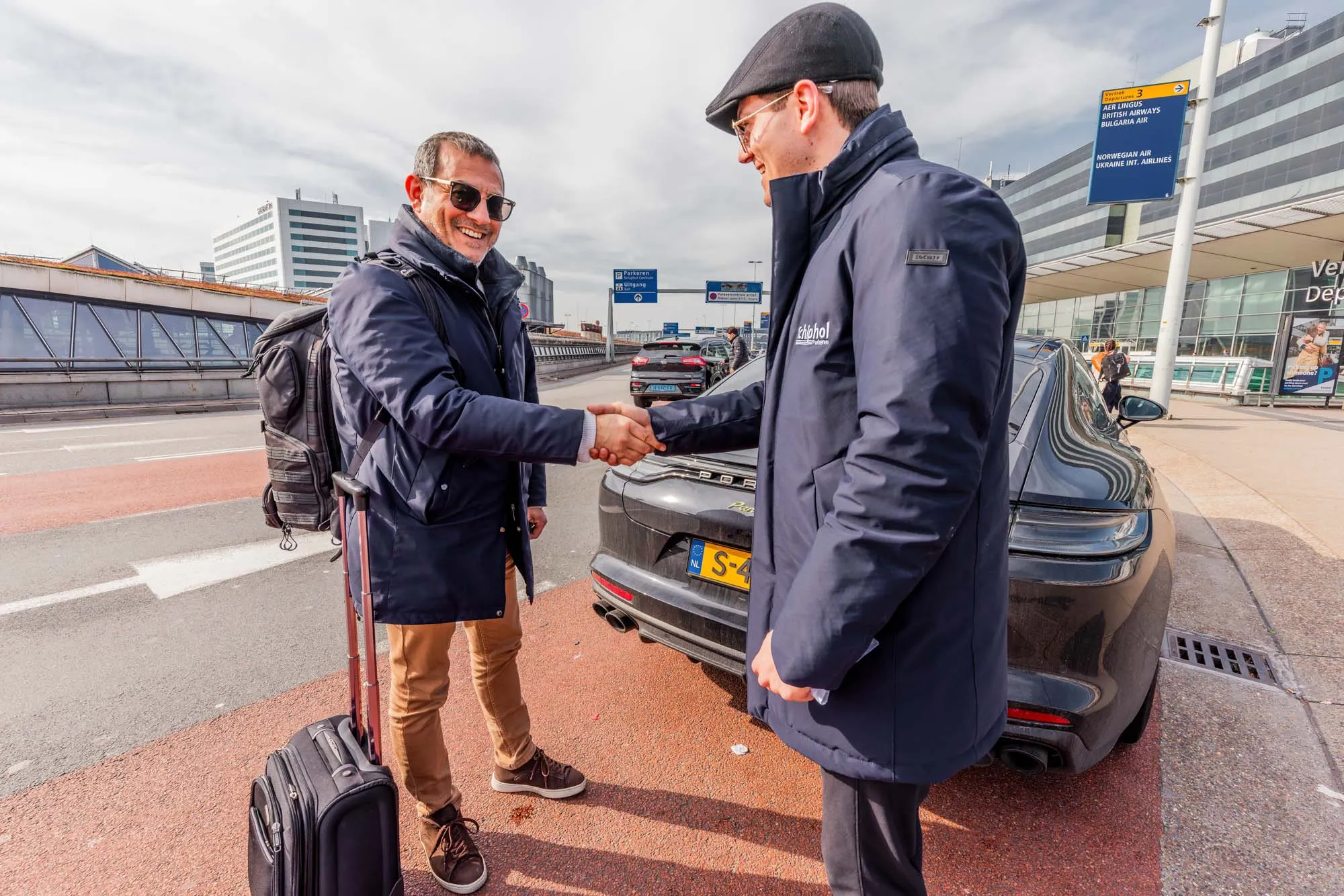 Two men shaking hands near a black car parked outside an airport terminal, one with luggage and a backpack.