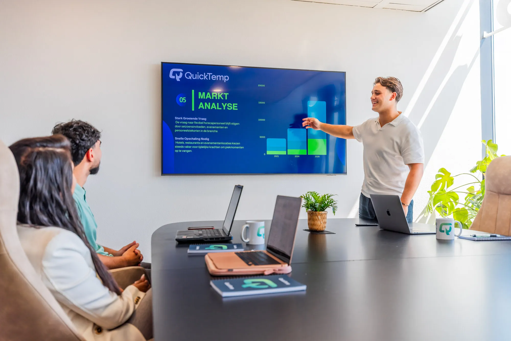 Man in white shirt presenting a market analysis slide to two seated colleagues in a bright office conference room.