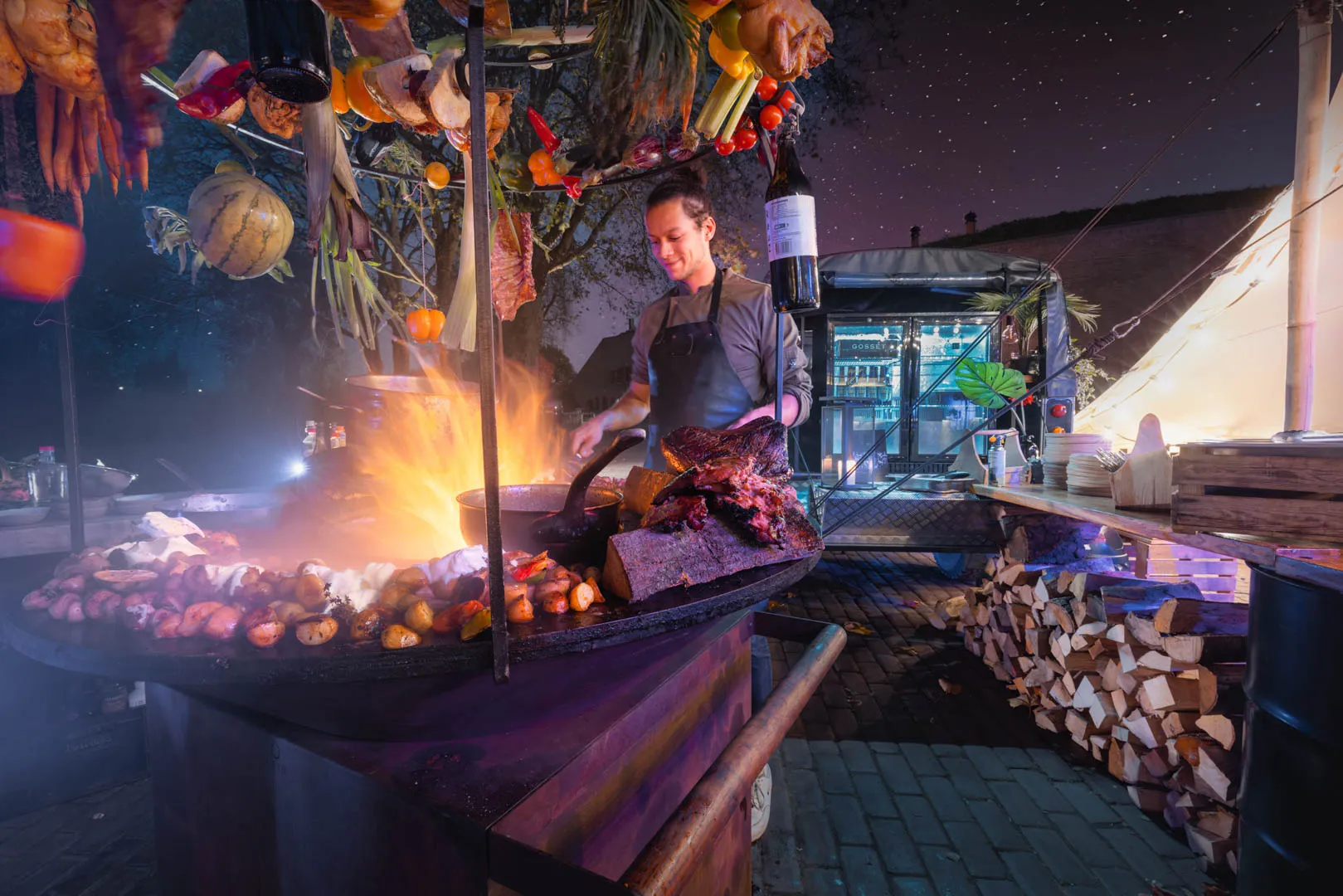 Chef cooking meat and sausages over an open flame grill with hanging vegetables and a wine bottle overhead at an outdoor nighttime food stall.