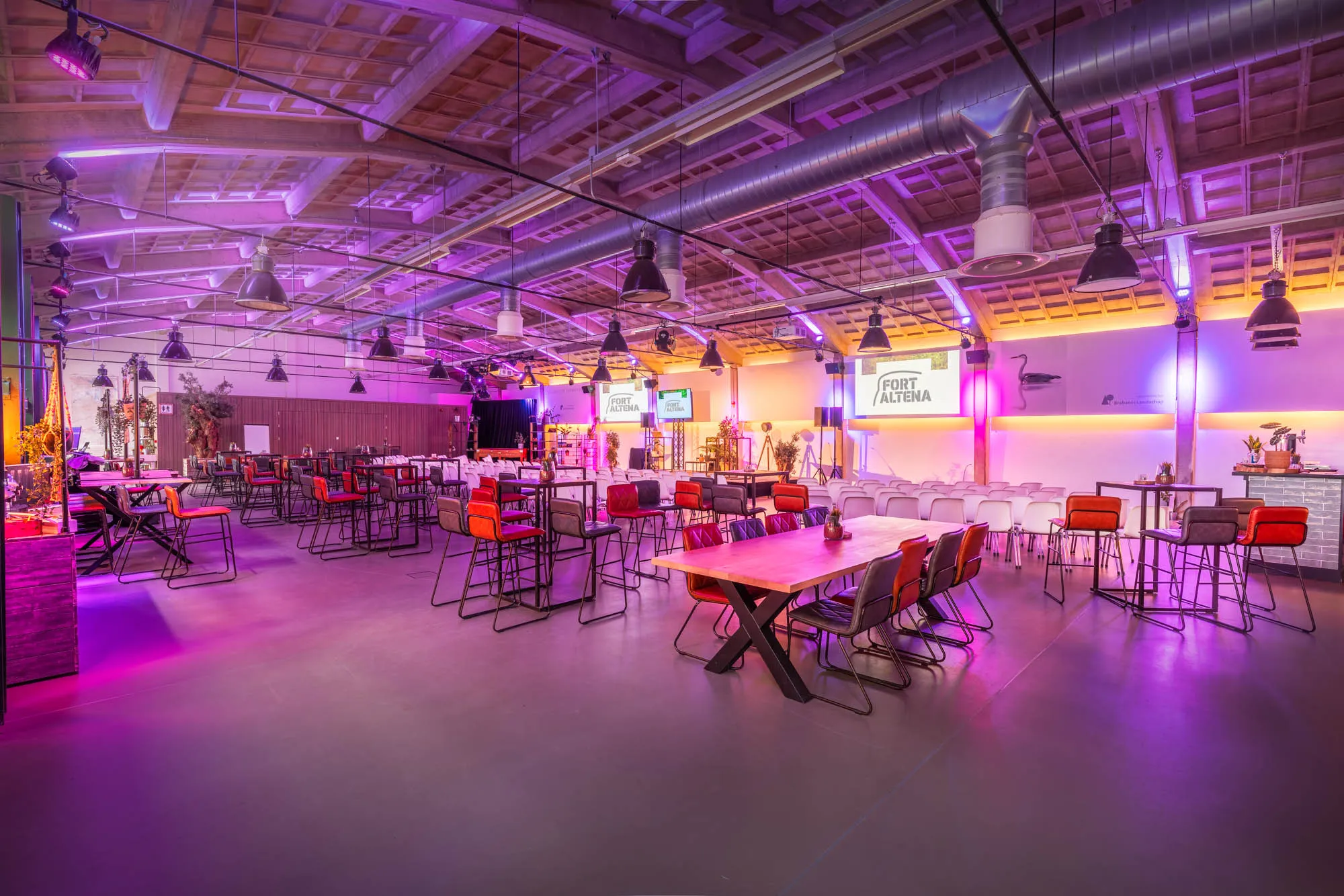 Spacious event hall with wooden ceiling, purple lighting, multiple tables and chairs, and screens displaying 'Fort Altena'.