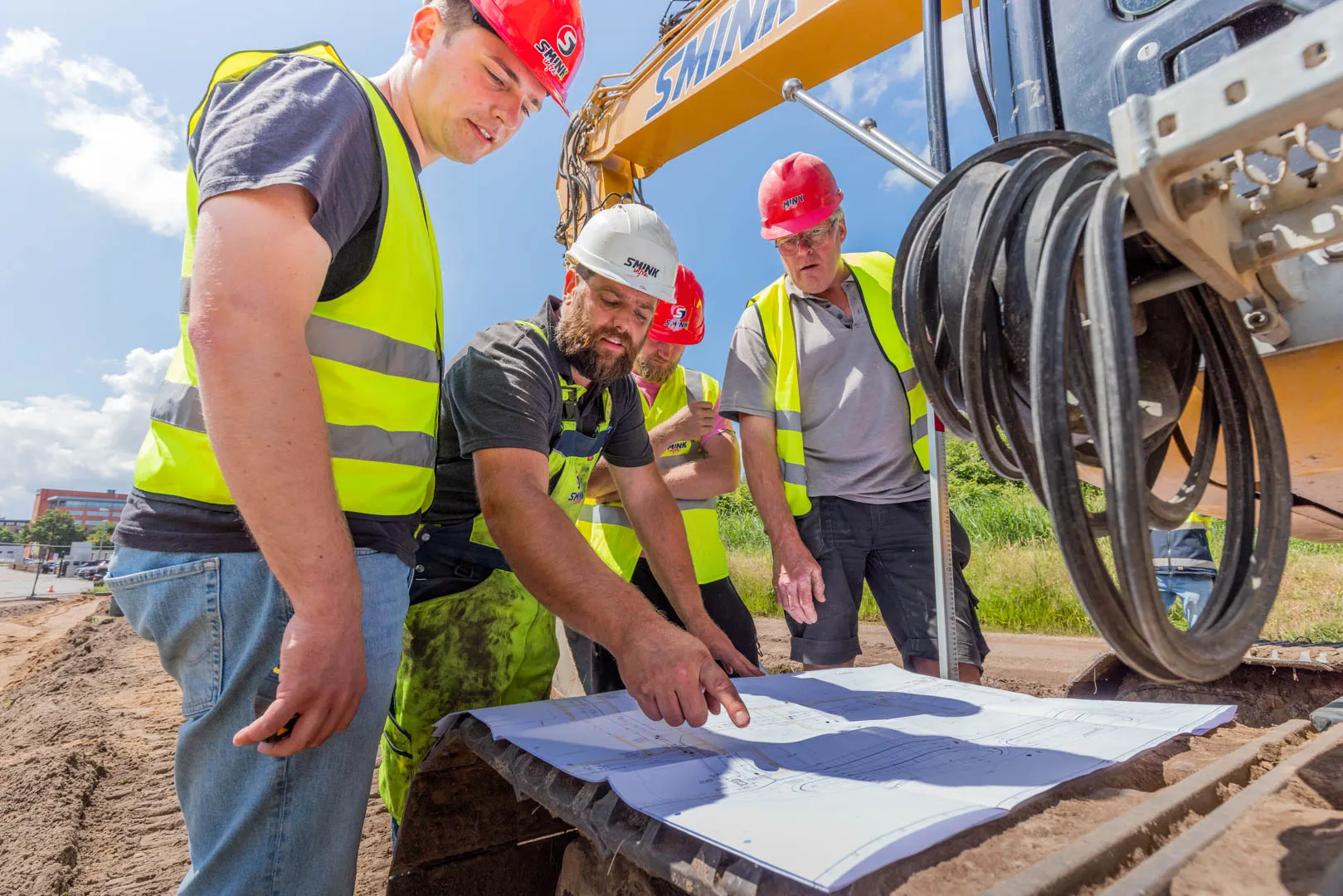 Four construction workers in safety vests and helmets examining blueprints on construction equipment under a sunny sky.