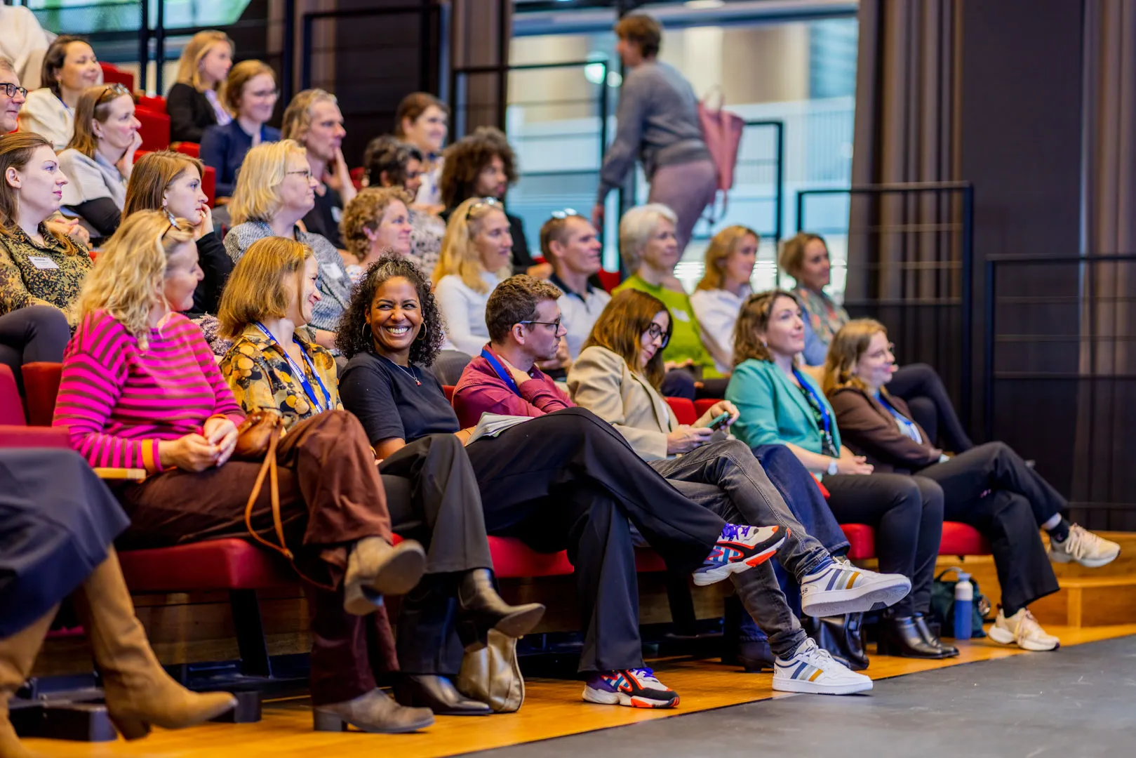 Audience seated in a conference room with red chairs, attentively watching and smiling during an event.