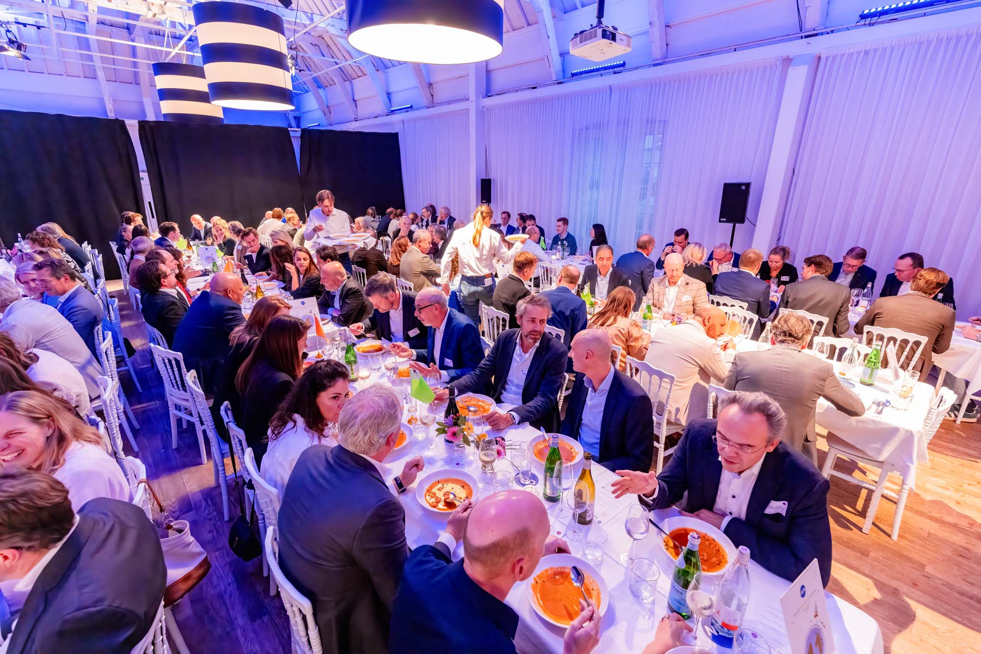 Two men in white shirts holding large arrow signs labeled 'HOPE' and 'NaaS' inside a spacious, industrial-style event space with pink and purple lighting and groups of people socializing.