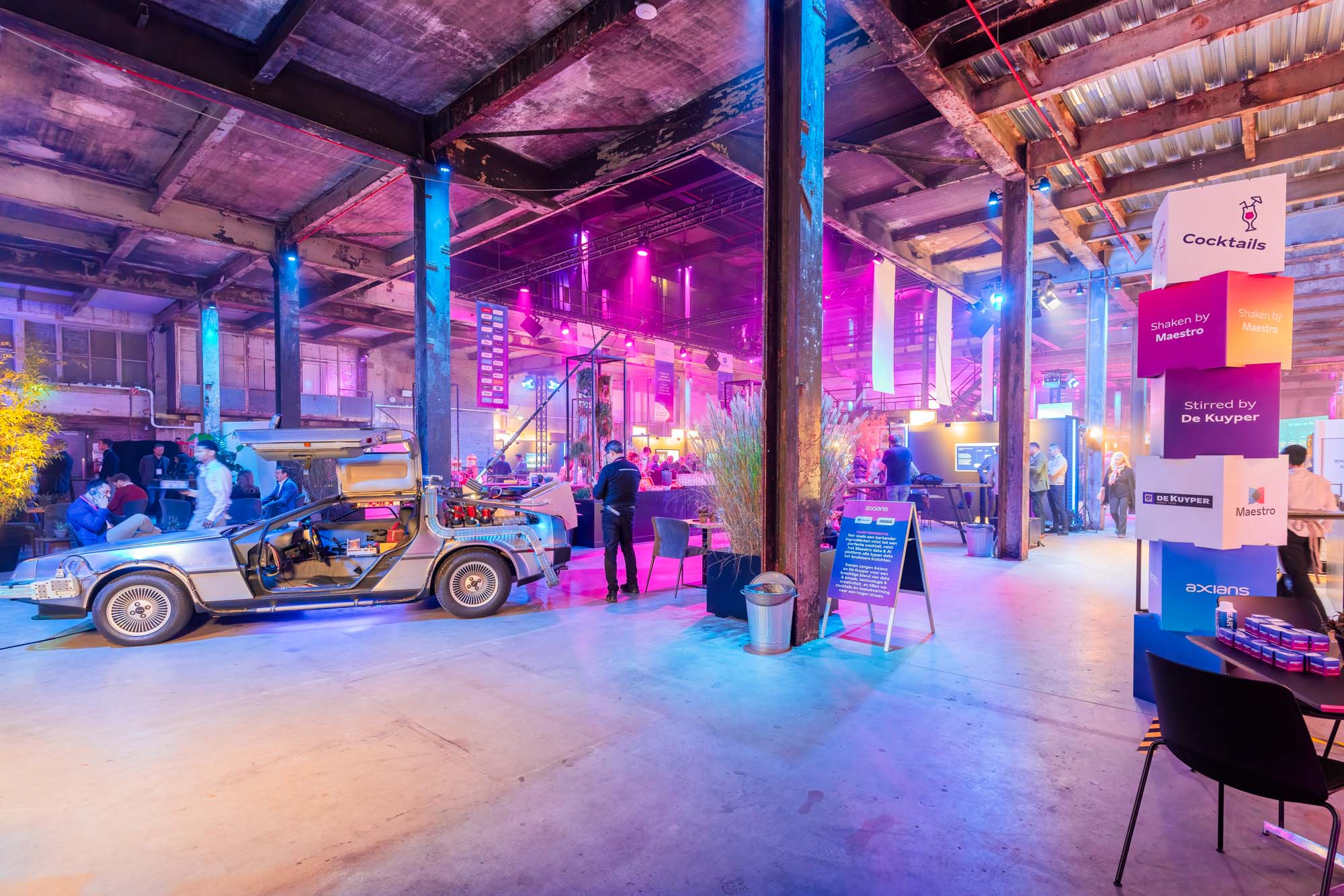 Two men in white shirts holding large arrow signs labeled 'HOPE' and 'NaaS' inside a spacious, industrial-style event space with pink and purple lighting and groups of people socializing.