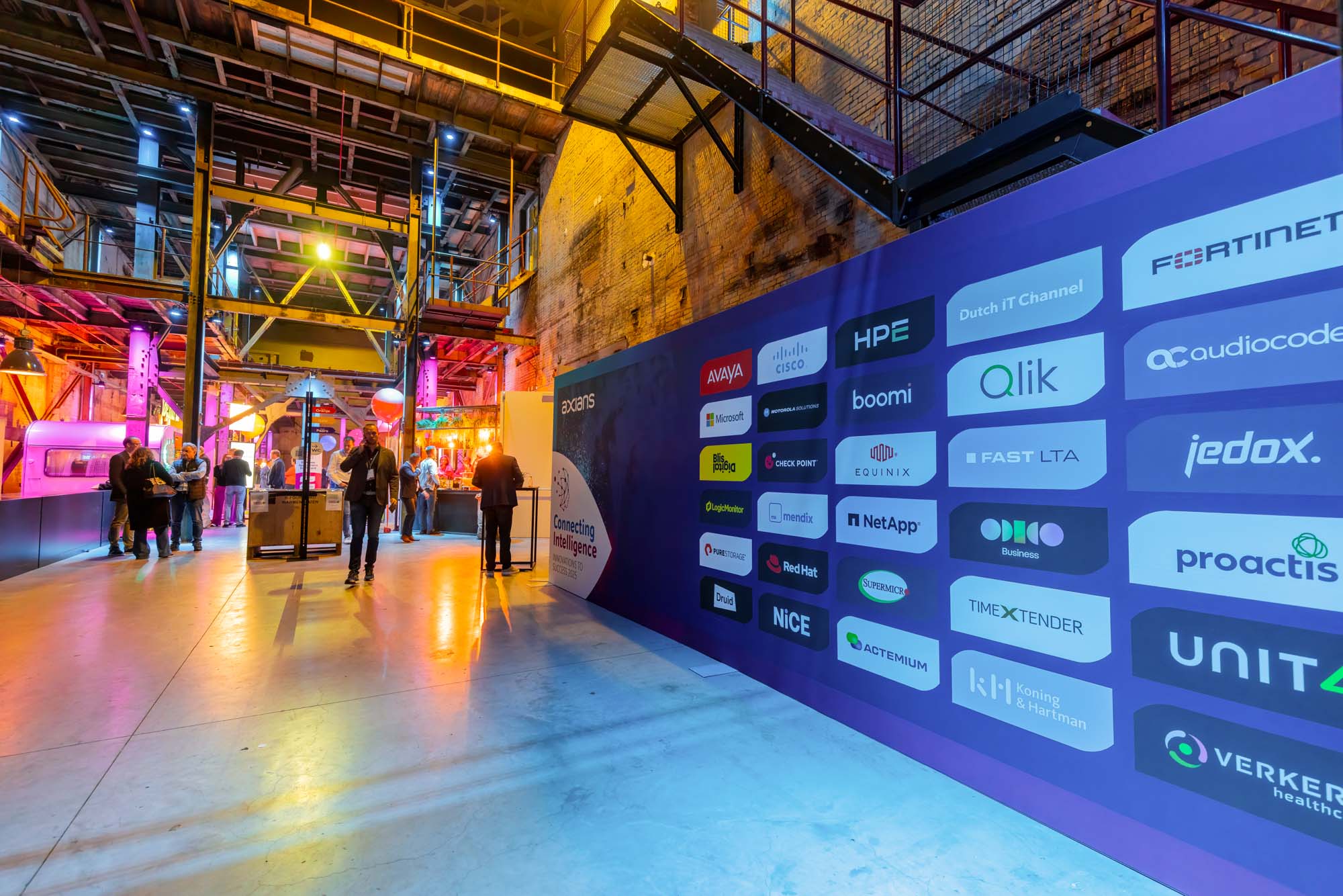 Two men in white shirts holding large arrow signs labeled 'HOPE' and 'NaaS' inside a spacious, industrial-style event space with pink and purple lighting and groups of people socializing.