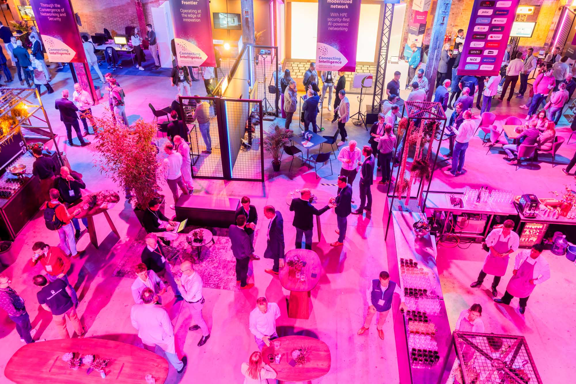 Two men in white shirts holding large arrow signs labeled 'HOPE' and 'NaaS' inside a spacious, industrial-style event space with pink and purple lighting and groups of people socializing.