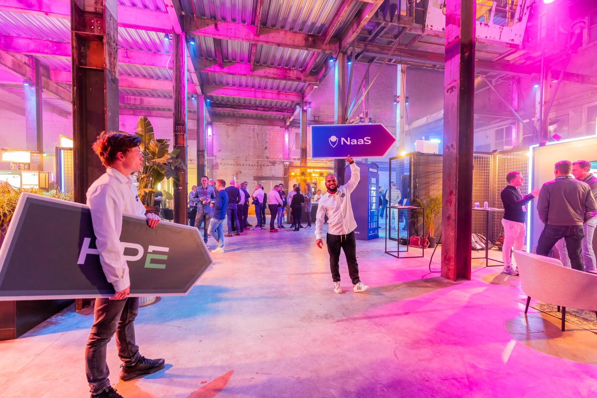 Two men in white shirts holding large arrow signs labeled 'HOPE' and 'NaaS' inside a spacious, industrial-style event space with pink and purple lighting and groups of people socializing.