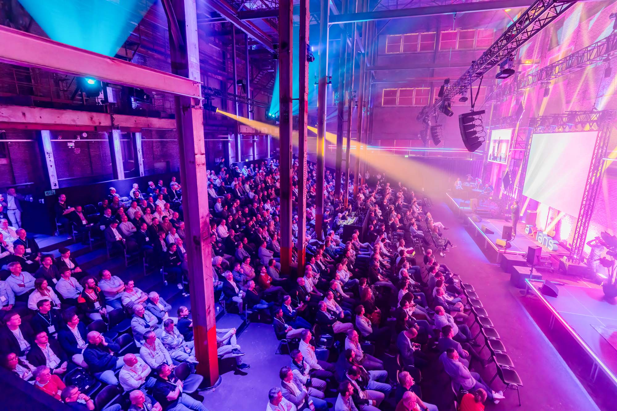 Two men in white shirts holding large arrow signs labeled 'HOPE' and 'NaaS' inside a spacious, industrial-style event space with pink and purple lighting and groups of people socializing.